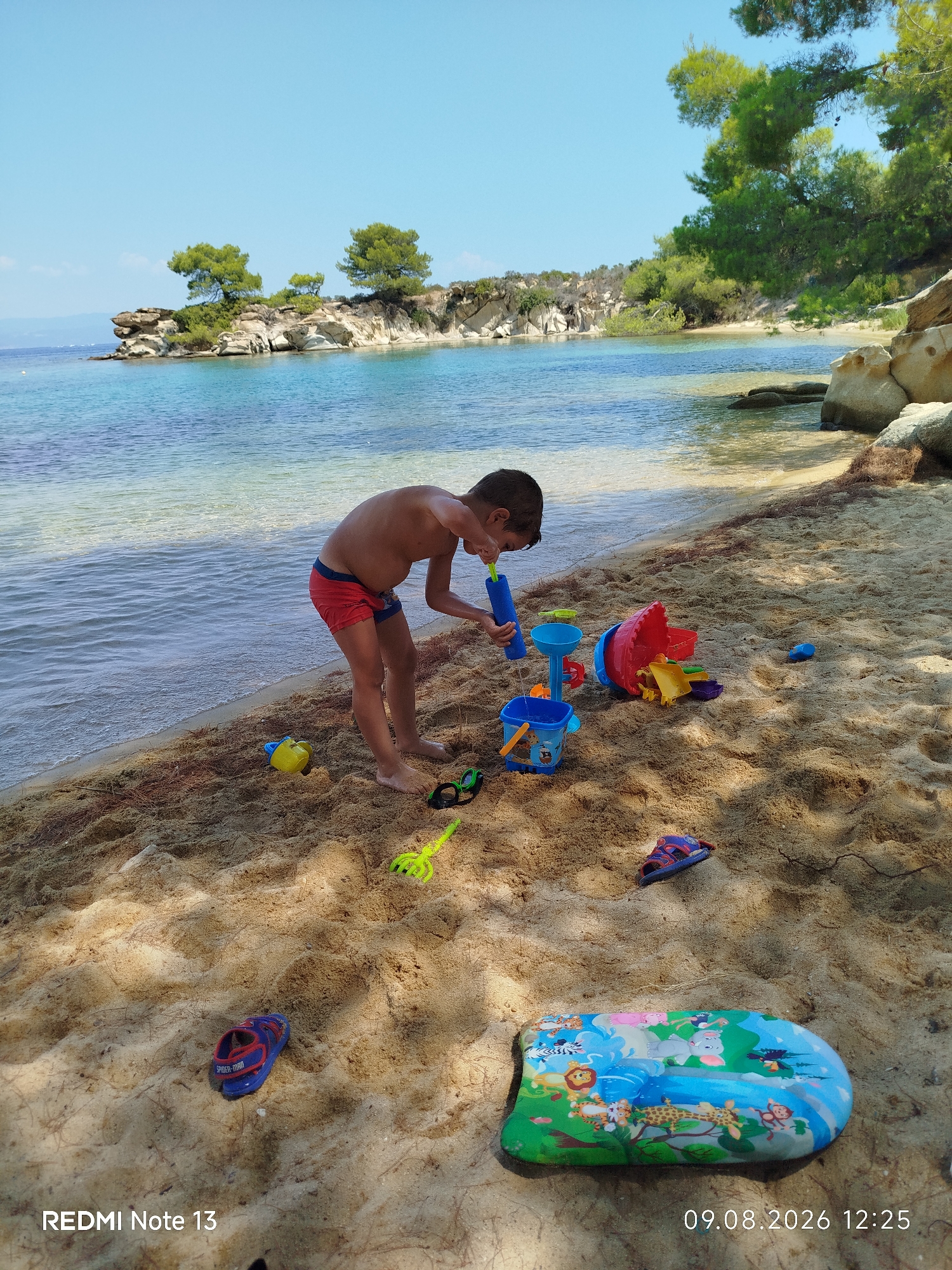 A young boy is playing with sand toys on a sandy beach in Sithonia Municipal Unit, Greece. The boy is wearing red swim trunks and is bent over a blue bucket, holding a blue water squirter. Scattered around him are various other colorful sand toys, including a red pail with yellow slides, a yellow bucket, and a green plastic rake. To the left of the boy, the clear blue water of the Aegean Sea laps gently against the shore. In the background, there are rocky outcroppings with sparse vegetation, and the characteristic pine trees of the region. The sky is clear and blue. In the foreground, a pair of child's sandals with a Spider-Man design and an inflatable water toy featuring cartoon animals are visible on the sand. The timestamp "09.08.2026 12:25" and the text "REDMI Note 13" are overlaid on the bottom of the image.