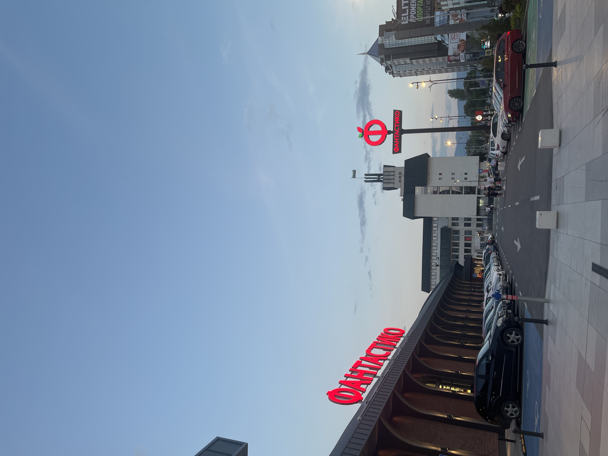 The image depicts a view of the Fantastico supermarket in Plovdiv, Bulgaria. The red "ФАНТАСТИКО" sign is prominently displayed on the building's facade. A row of parked cars lines the right side of the image. Across the street stands another Fantastico sign with an apple logo on a pole. Buildings, including a taller modern structure, are visible in the background, along with streetlights and some pedestrian activity. The sky is clear blue.