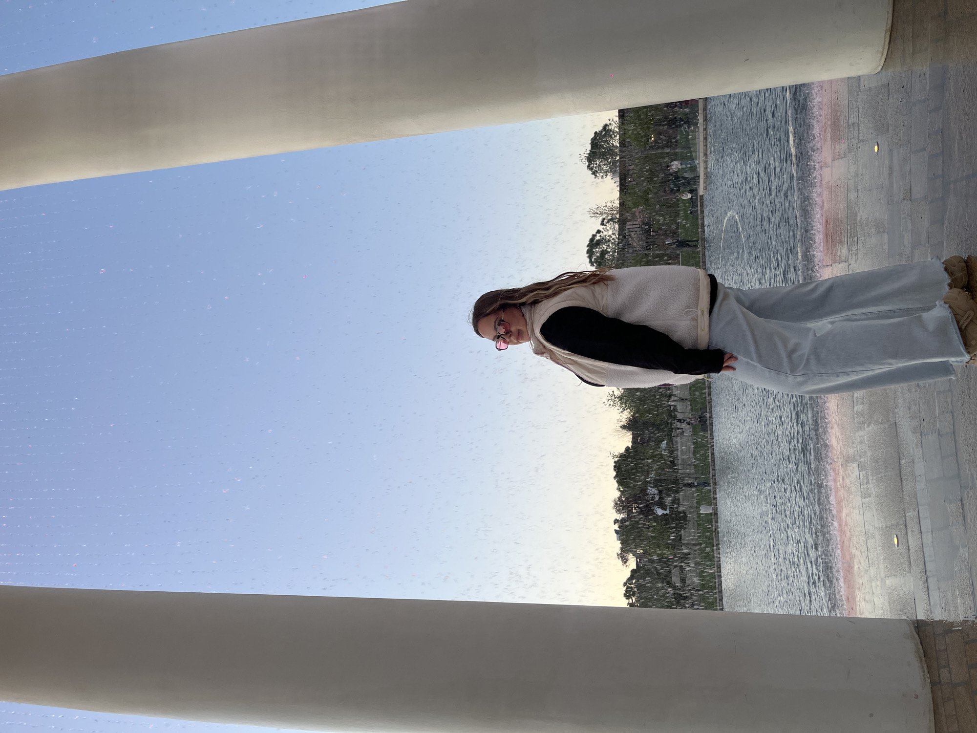A young woman with light brown hair and pink glasses stands facing left in an outdoor setting. She wears a white sherpa vest over a black shirt and light blue jeans, paired with tan-colored shoes. The woman appears to be standing beneath a structure featuring a prominent white pillar. A body of water extends horizontally behind her, lined by trees and greenery. Light-colored pavement is visible on the right side of the frame. The sky is light blue, suggesting daytime. Small pink dots are scattered across the image, resembling water droplets on a lens. The location is the Krasnodar Musical Fountain.