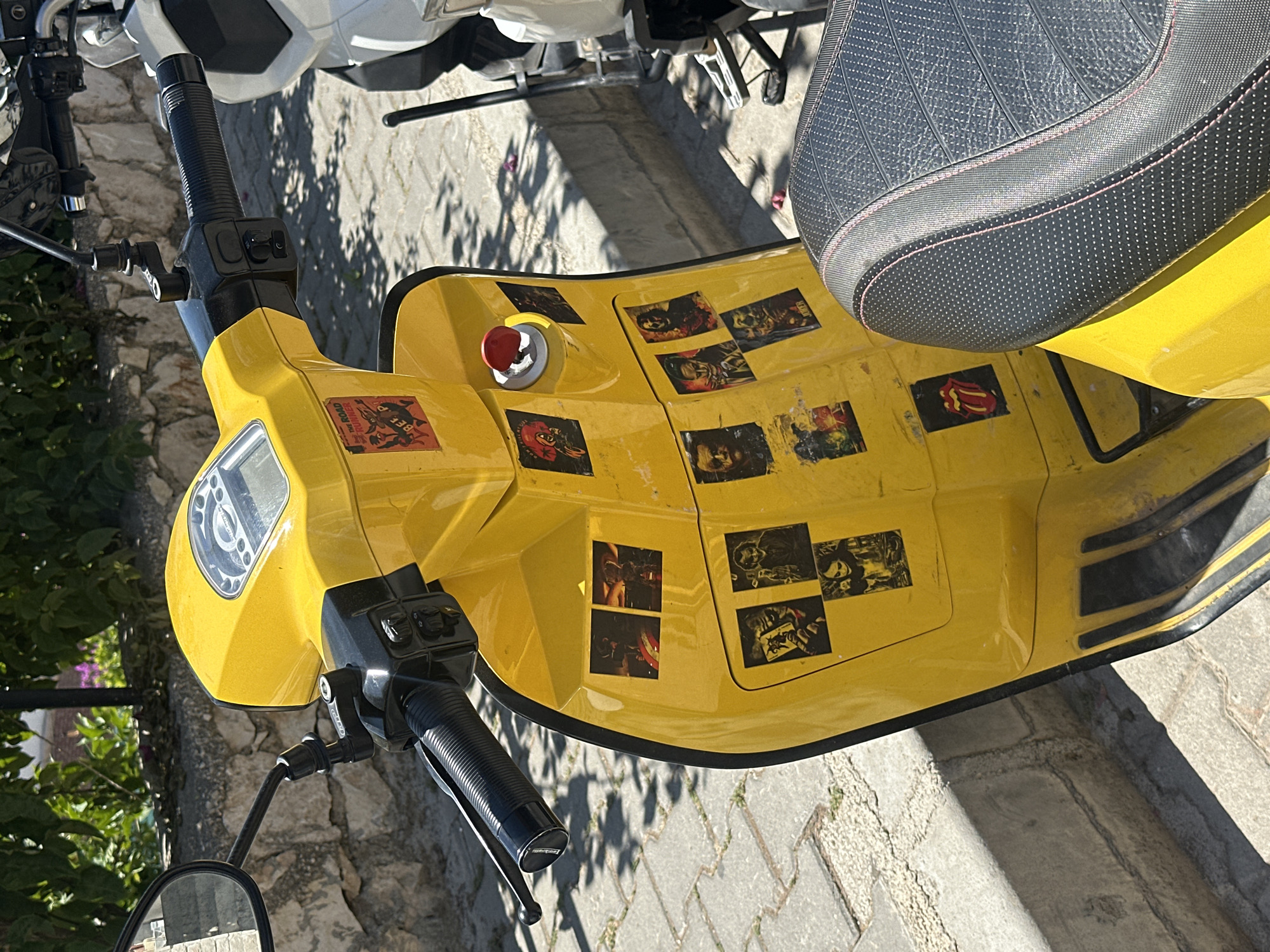 The image captures a yellow scooter parked outdoors in Kaş, Turkey, under bright daylight. The primary subject is the front section of the scooter, viewed from a high angle, showcasing its vibrant yellow body, a black textured seat with red stitching, and black handlebars.
The scooter's leg shield and footboard area are notably adorned with numerous stickers. Visible text includes "THE ROAD RUNNER" on a sticker towards the front left, and a red and yellow sticker nearby that appears to read "BEE P" or similar. Further back, a recognizable red Rolling Stones tongue logo is present. Many other small, mostly monochrome or dark-toned stickers depict various graphic designs, possibly band logos or album art, though they are largely indistinct. A red ignition button or cap is also visible on the yellow panel.
The setting features light-colored paved ground beneath the scooter, possibly cobblestones or pavers, consistent with a sidewalk or street. Long shadows cast across the ground indicate clear, sunny weather, suggesting it is either morning or late afternoon. Green foliage and bushes are visible in the background to the left, adding a natural element to the scene. The scooter is stationary, and no people or specific activity are visible.