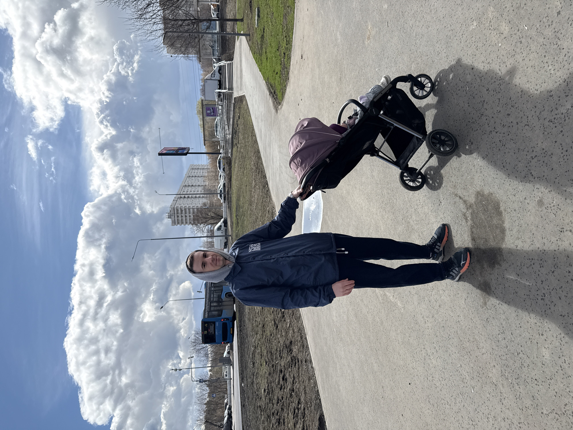 A man in a blue jacket and dark pants stands on a concrete path next to a baby stroller, facing the camera. The stroller has a purple hood and appears to be carrying a child. The man wears a gray hooded sweatshirt underneath his jacket and dark sneakers. The path runs alongside a grassy area with some trees. In the background, there is a road with parked cars and a blue bus. Further back, there are residential buildings. The sky is partly cloudy. The setting is consistent with a park area in a city, likely Moscow, Russia.