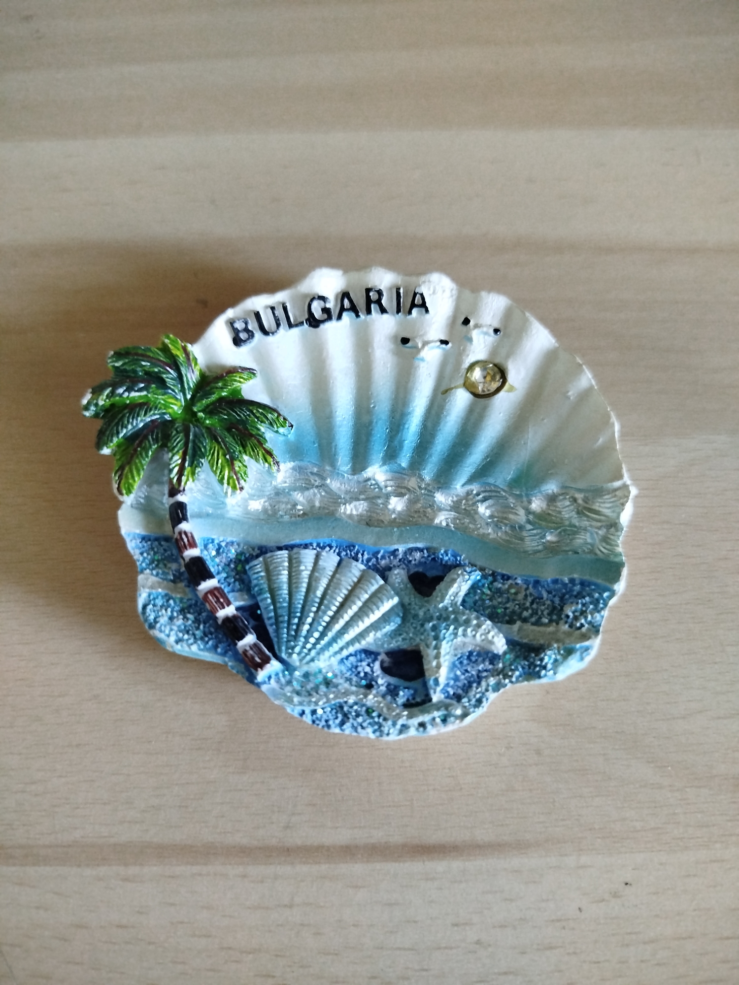 A close-up shot shows a souvenir magnet of Bulgaria resting on a light brown wooden surface. The magnet is shaped like a scallop shell and decorated with a coastal scene. The word "BULGARIA" is printed in capital letters at the top of the shell. The scene includes a palm tree on the left side with a black and brown segmented trunk. To the right of the tree, there is a depiction of a blue sea with white waves. In the bottom portion of the magnet, there's a fan-shaped seashell and a starfish nestled in blue glittery sand. A small gold-colored embellishment is positioned near the top of the shell. The souvenir suggests a beach theme, possibly representing Varna, Bulgaria, a coastal city known for its beaches.