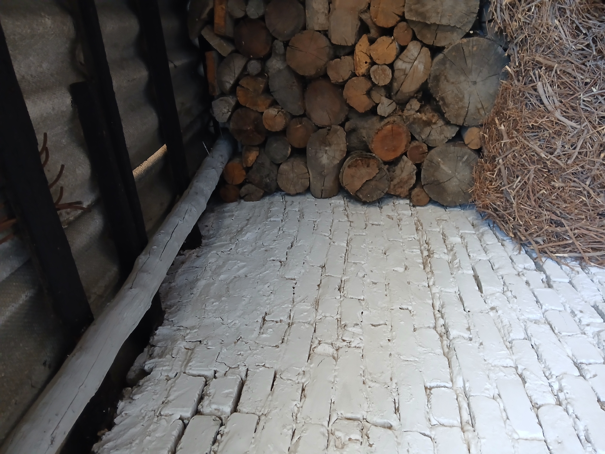 The image depicts a corner of an outdoor storage area, possibly a shed or lean-to, located in Koktal, Kazakhstan. The left side of the image is dominated by a white-washed brick wall, showing signs of wear and cracking. A rough-hewn wooden beam runs horizontally across the top of the wall. The right side of the image is filled with a neatly stacked pile of cut firewood of various sizes. The firewood is a mix of light and dark brown tones, with visible wood grain and rings. In the foreground, beneath the firewood, lies a pile of dry, thin branches and twigs, likely used for kindling or as bedding material. The roof structure above is visible, consisting of corrugated material supported by dark wooden beams. The overall scene suggests a functional outdoor space for storing fuel.