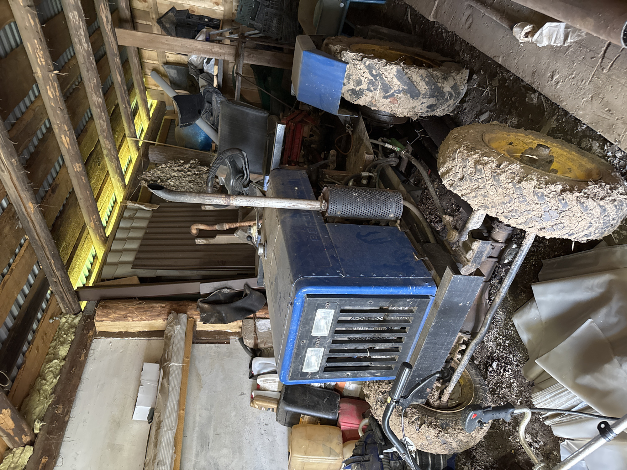 The image shows a blue and black tractor parked inside a rustic, wooden structure that appears to be a barn or shed. The tractor is heavily caked in mud, particularly its tires, suggesting it has been used in a muddy environment. The interior of the structure is cluttered with various items, including plastic containers, tools, what looks like insulation material, and assorted debris. The roof is made of exposed wooden beams and corrugated metal. The overall scene depicts a functional, albeit messy, agricultural or storage space in a rural setting. The location is specified as Бородулино, Russia. There are no people visible in the image.