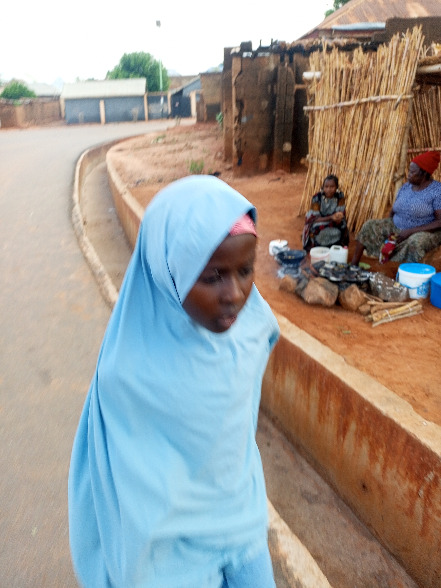 The image displays an outdoor scene within a semi-urban or rural residential setting in the city of Unknown, Unknown. In the immediate foreground, to the left, a young individual, likely female, is partially visible and in motion. This person wears a light blue full-body hijab with a pink band visible beneath it, and is looking downwards and slightly to the right.
To the left of the frame, a greyish, curved paved road or pathway is present, accompanied by a concrete curb and an open concrete drainage gutter. The ground immediately to the right of the gutter and extending across the midground is composed of reddish-brown dirt, which appears dry and uneven.
In the midground, on the right side of the frame, two adult women are seated directly on the reddish-brown earth. The woman on the left of this pair wears a patterned dress and a head covering, with her hands appearing engaged in an activity in her lap or on the ground. The woman on the right wears a blue patterned top, a patterned long skirt, and a red head covering, holding a white object in her lap. Between these two women, an open-air cooking setup is visible. It features several dark rocks arranged around a patch of dark ash, suggesting a recent fire. A dark-colored pot or bowl rests on some of these rocks. Adjacent to the cooking area are stacks of firewood and various plastic containers, including two white rectangular containers, one white bucket with a blue lid, and one entirely blue bucket.
The background comprises several structures. Directly behind the seated women is a rustic screen or wall made of numerous vertical wooden poles or reeds. Beyond this, a weathered building constructed from mud-bricks or similar traditional materials with a visible roof (possibly corrugated iron or thatched) is present. Further in the distance, additional structures and sparse green vegetation, including trees, are discernible under a bright, diffused sky.