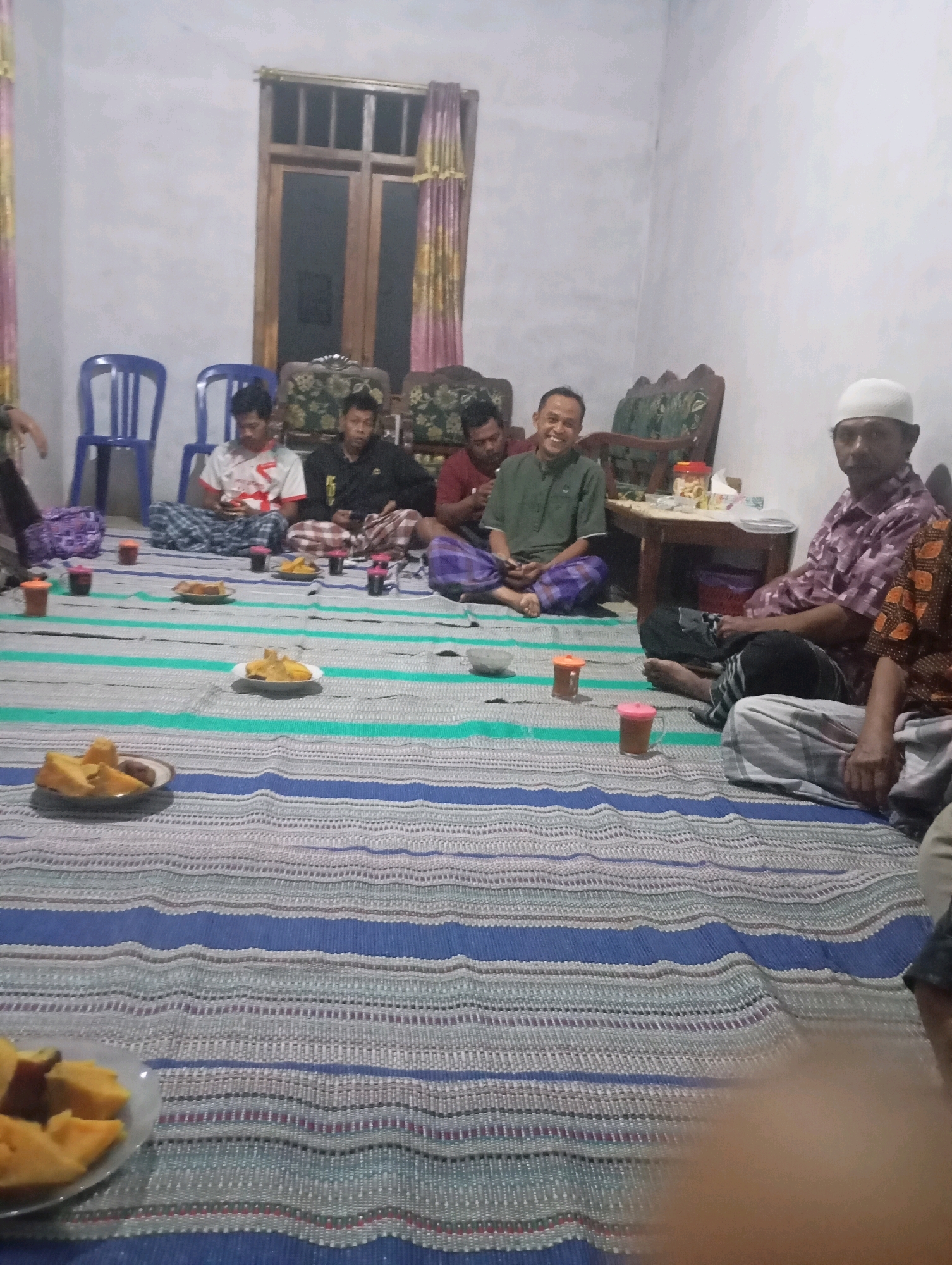 A group of men are gathered in a room, possibly a living room, in Ketepung, Indonesia. They are seated on a mat spread on the floor, facing a low table. Several plates of food, likely snacks or appetizers, are visible on the mat and table, along with cups of drinks. Some of the men are dressed in traditional Indonesian attire, including sarongs and peci hats. They appear to be engaged in conversation or a communal meal. The room's decor includes wooden furniture, curtains, and a window with wooden blinds. The lighting is dim, suggesting it might be evening or the room is not well-lit. The overall atmosphere seems relaxed and informal.