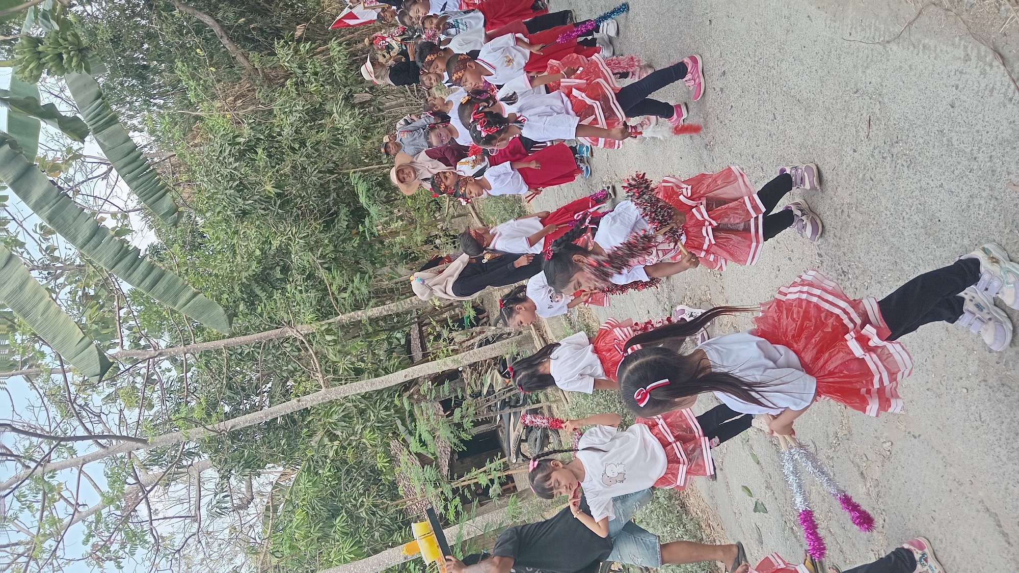 A procession of children in red and white costumes walks down a rural dirt road in Kedungwaru, Indonesia. The children, wearing white t-shirts and red skirts, appear to be performing or celebrating. Some are holding decorative batons. Adults are visible in the background, with one woman in a headscarf and another wearing a straw hat. Lush greenery and trees line the road, suggesting a tropical or subtropical environment. The sky is overcast.