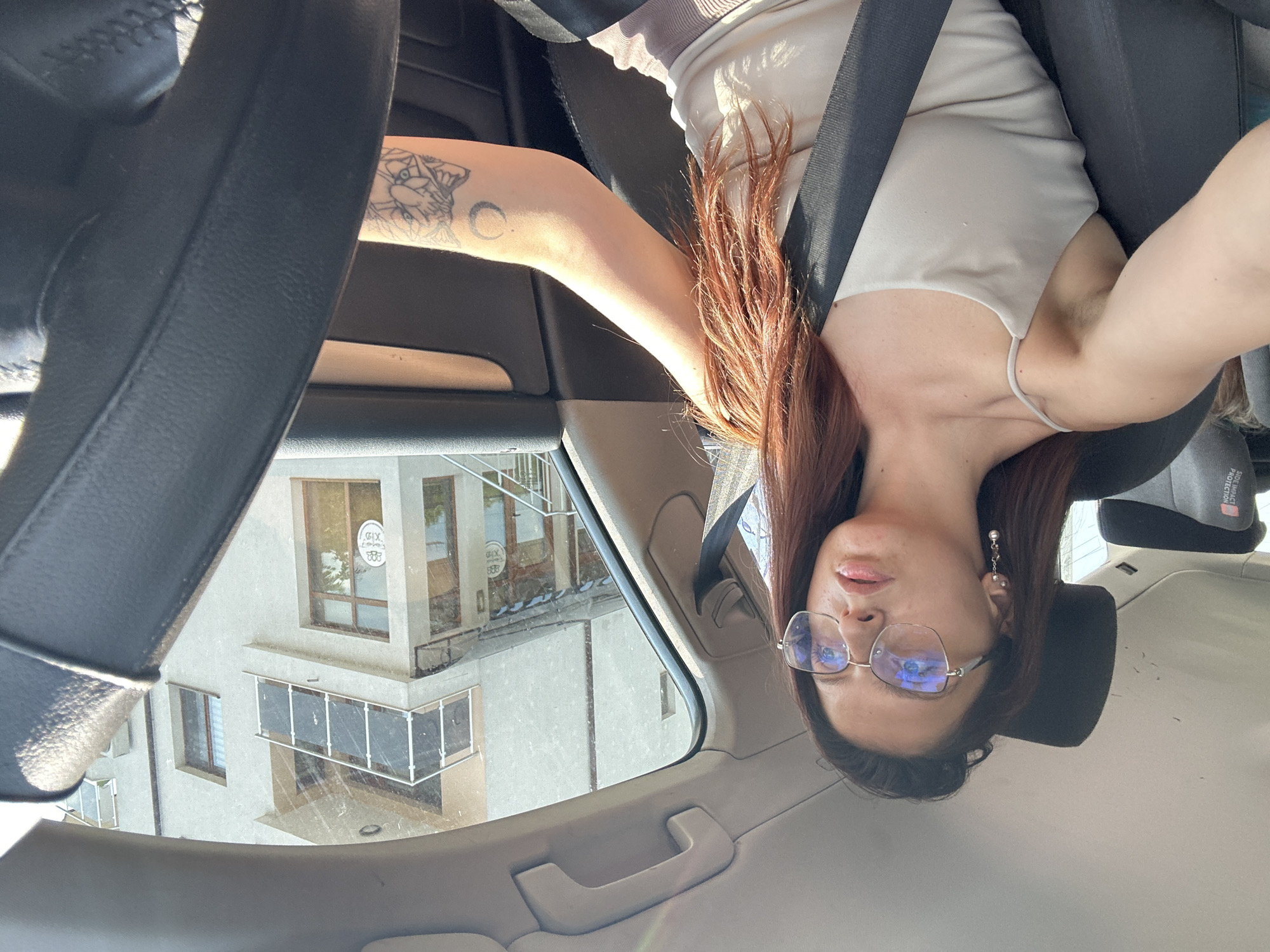 A young woman with long, auburn hair and clear-rimmed glasses is seen from the chest up, sitting in the driver's seat of a car, driving on a sunny day. She is wearing a light-colored tank top and has a seatbelt fastened across her body. Her right arm, which rests on the steering wheel, has a tattoo of a moon and a design of two figures. In the background, through the car window, a light-colored building with balconies and windows is visible, characteristic of urban architecture in Tryavna, Bulgaria. The sun is casting shadows and highlighting dust particles in the air, visible on the car's interior surfaces. The overall scene captures a moment of everyday life in a Bulgarian city.