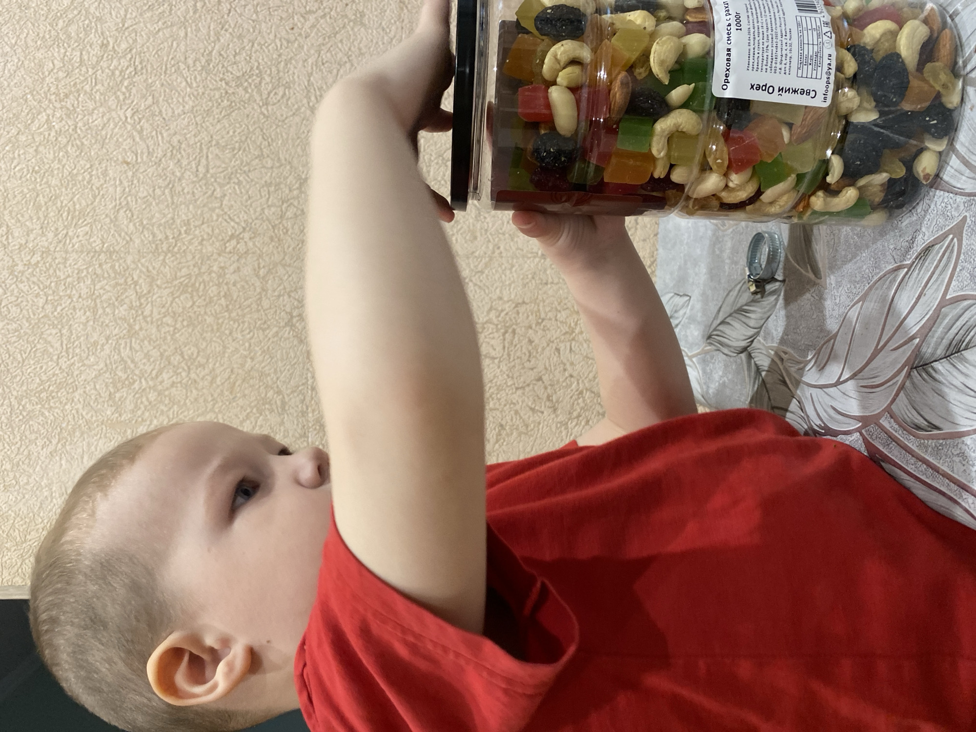 A child in a red shirt is holding a large plastic jar of mixed nuts and dried fruit. The jar contains cashews, almonds, raisins, and candied fruit pieces. The label on the jar reads "Ореховая смесь с рахат 1000г Свежий Орех". The label also shows "km 10:32, Moscow". The background shows a textured wall and a table or surface with a patterned cover.