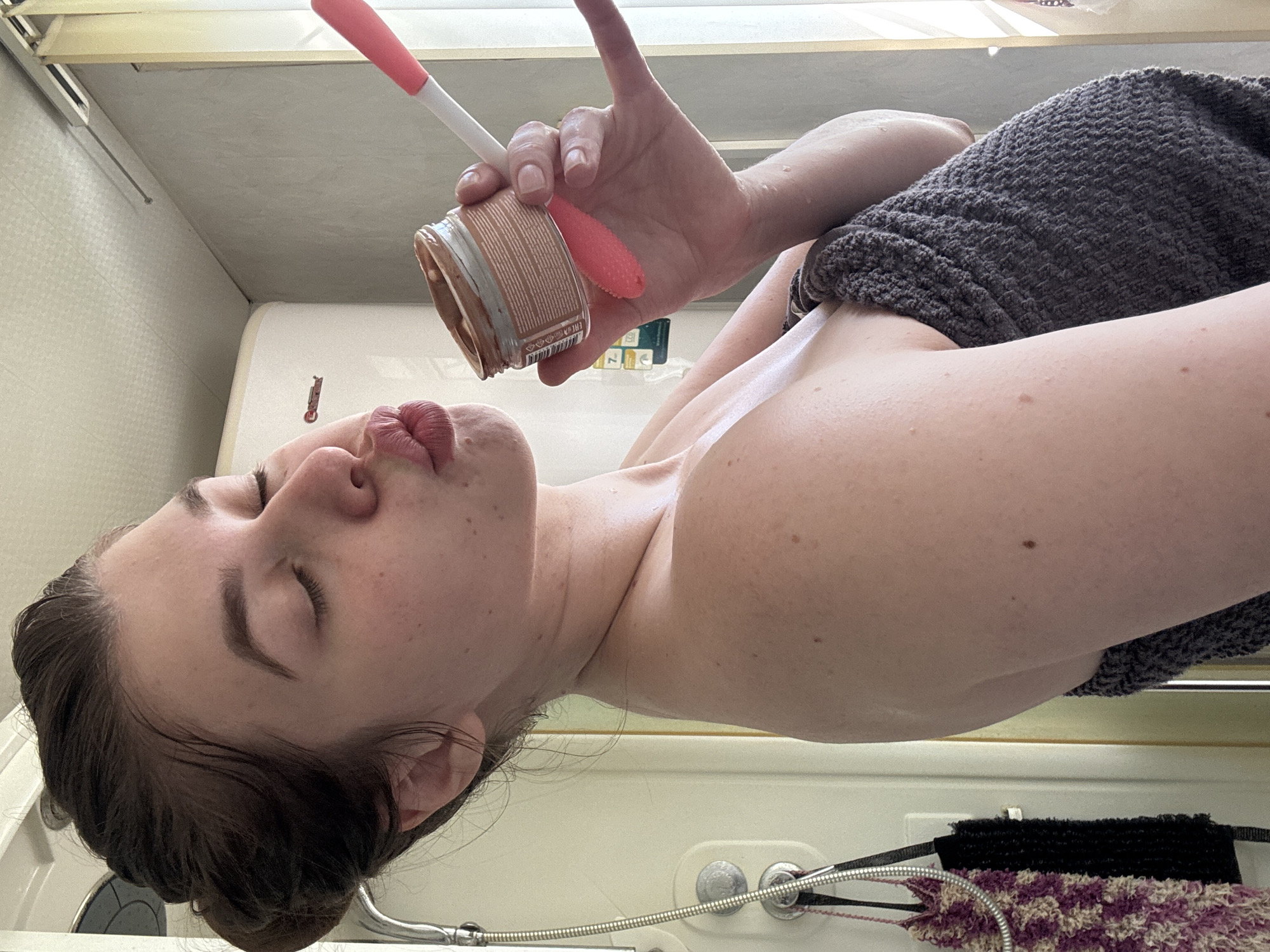 A woman is in a bathroom, holding a jar of brown substance with a red applicator. She is puckering her lips towards the jar. She has a gray towel draped over her shoulder. The background includes a shower and bathroom fixtures. There is no evidence to support that this image was taken in Шевлягино, Russia.