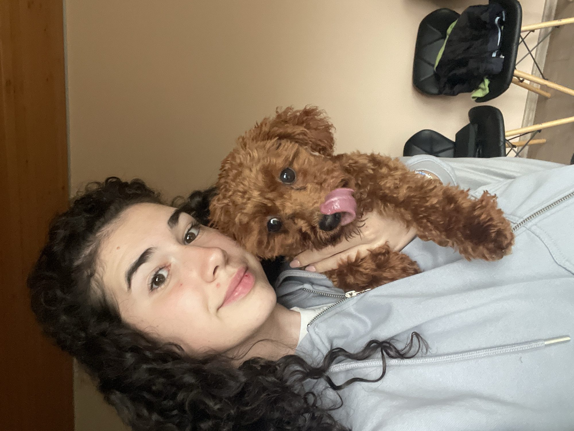A close-up, horizontally oriented selfie captures a young woman and a small dog in an indoor setting. The woman, positioned on the left, has dark, curly hair and light brown eyes, looking directly at the camera with a gentle smile. She wears a light grey hooded zipper jacket. Resting on or near her is a small, fluffy, reddish-brown dog, resembling a poodle, which is looking towards the viewer with its tongue playfully extended.
The background consists primarily of a plain, light beige wall. To the far left, a wooden door frame or piece of furniture is partially visible. In the upper right section, two black chairs with wooden legs are seen; one appears upright, and the other is either overturned or leaning. The scene suggests a casual, intimate moment between a pet owner and her dog, exuding a sense of warmth and affection. The lighting indicates an indoor environment, with no specific time of day or weather cues discernible. There is no visible text within the image. The setting is likely a home, potentially in Sofia, Bulgaria.