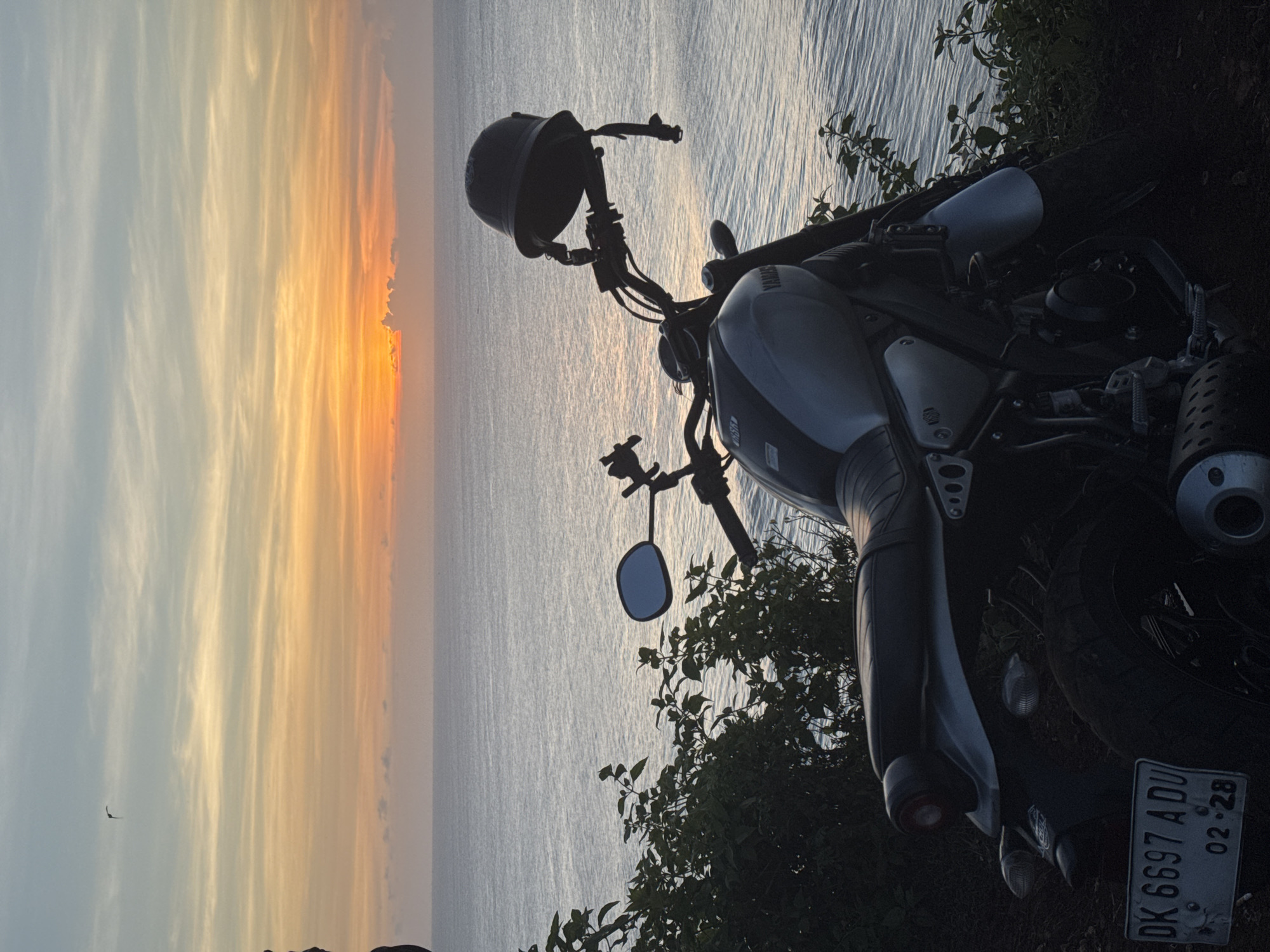 A motorcycle is parked on a hill overlooking the ocean at sunset. The sky is filled with shades of yellow and orange. The motorcycle has a helmet resting on the handlebars. The license plate reads "DK 6697 ADU" and "02·28." Green foliage surrounds the motorcycle. A small bird is visible in the sky. Location: Unknown, Unknown.