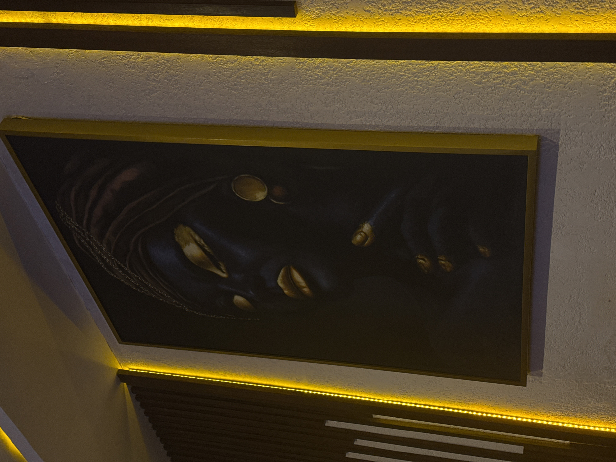 This image captures an interior wall decorated with contemporary lighting and artwork, likely in an establishment in North Kuta, Indonesia. The dominant feature is a large, rectangular framed artwork, set against a light-colored, textured wall. The frame of the artwork is a bright golden-yellow. The artwork itself is dark, possibly black or deep blue, and features abstract or stylized elements in gold or bronze, which faintly resemble facial features like eyes and lips, along with what could be braided hair or chains.
The lighting plays a crucial role in the ambiance. Three distinct lines of warm, yellow LED strip lights are visible. One strip is positioned above the framed artwork, illuminating the textured wall above it. A second strip runs along the top edge of the artwork. The third strip is located below the artwork, casting light downwards onto the textured wall and a decorative panel of horizontal, dark wooden slats. The arrangement of the lighting creates a soft, inviting glow, highlighting the textures of the wall and the artwork.
The overall scene suggests an evening or nighttime setting, as the interior relies entirely on artificial lighting. There are no people visible, nor any specific activity taking place; the image focuses on the decorative elements and the atmosphere created by the lighting. No discernible text is present in the image. The style of decor, with textured walls, warm lighting, and dark, abstract art, points towards a modern and sophisticated interior design.