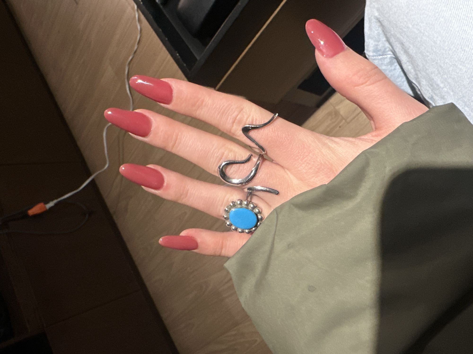 A close-up photograph shows a hand with long, almond-shaped fingernails painted a glossy rose-brown. The hand is adorned with two rings: a silver ring with a wavy, abstract design on the middle finger, and a silver ring with a turquoise stone surrounded by small silver beads on the ring finger. The hand is positioned against a background that includes a light-colored wooden surface, a section of what appears to be a dark black box, a light blue denim fabric, and a green jacket or garment partially covering the hand. A white cable with an orange connector is also visible in the background. The location is not discernible from the provided image.