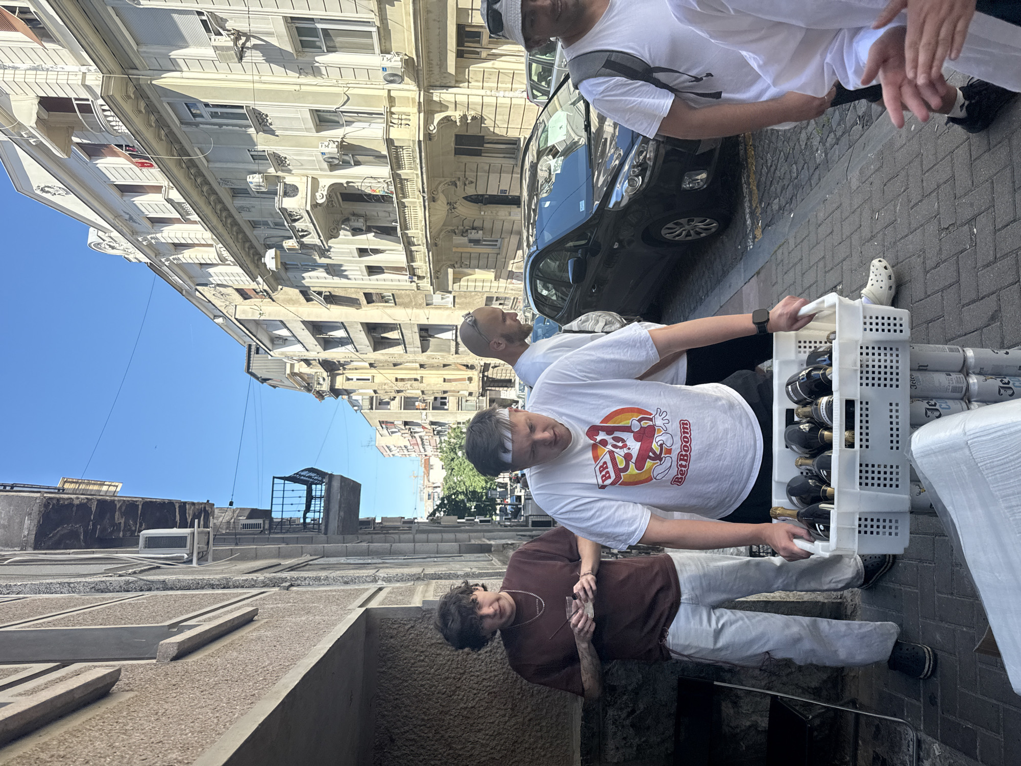 The image depicts an outdoor scene, presumably a street in Belgrade, Serbia. The architecture suggests an urban setting. Four men are visible, engaged in what appears to be the delivery of beverages. One man is wearing a white shirt with a cartoon graphic and a white headband. He is holding a white plastic crate filled with glass bottles, potentially wine or champagne, and aluminum cans. Another man wears a brown shirt. Other men wear white t-shirts. A black car is parked nearby. The street is paved with rectangular blocks. Buildings line the street, some with decorative facades and multiple windows. The sky is blue.