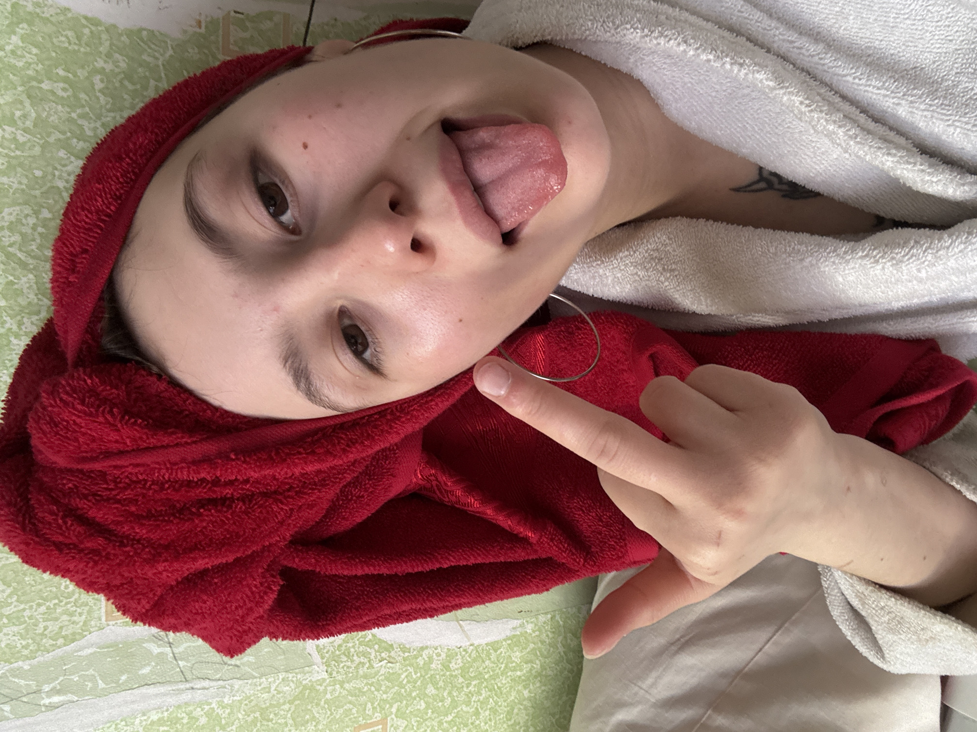The image displays a close-up, frontal portrait of a young woman in an indoor setting in the city of Фоки, Russia. She has fair skin and dark brown eyes. Her head is wrapped in a bright red terrycloth towel, and she is wearing a light-colored terrycloth bathrobe. A large silver hoop earring is visible on her left ear. She is making a defiant gesture, extending her tongue from her open mouth and raising her left hand with the middle finger extended upwards towards the camera. Multiple linear scars are visible on her right wrist and forearm. A dark tattoo, possibly tribal in style, is partially visible on her upper chest beneath the bathrobe. The background consists of green, textured wallpaper showing signs of wear and tear, with a section of light-colored fabric, likely bedding, to the left.