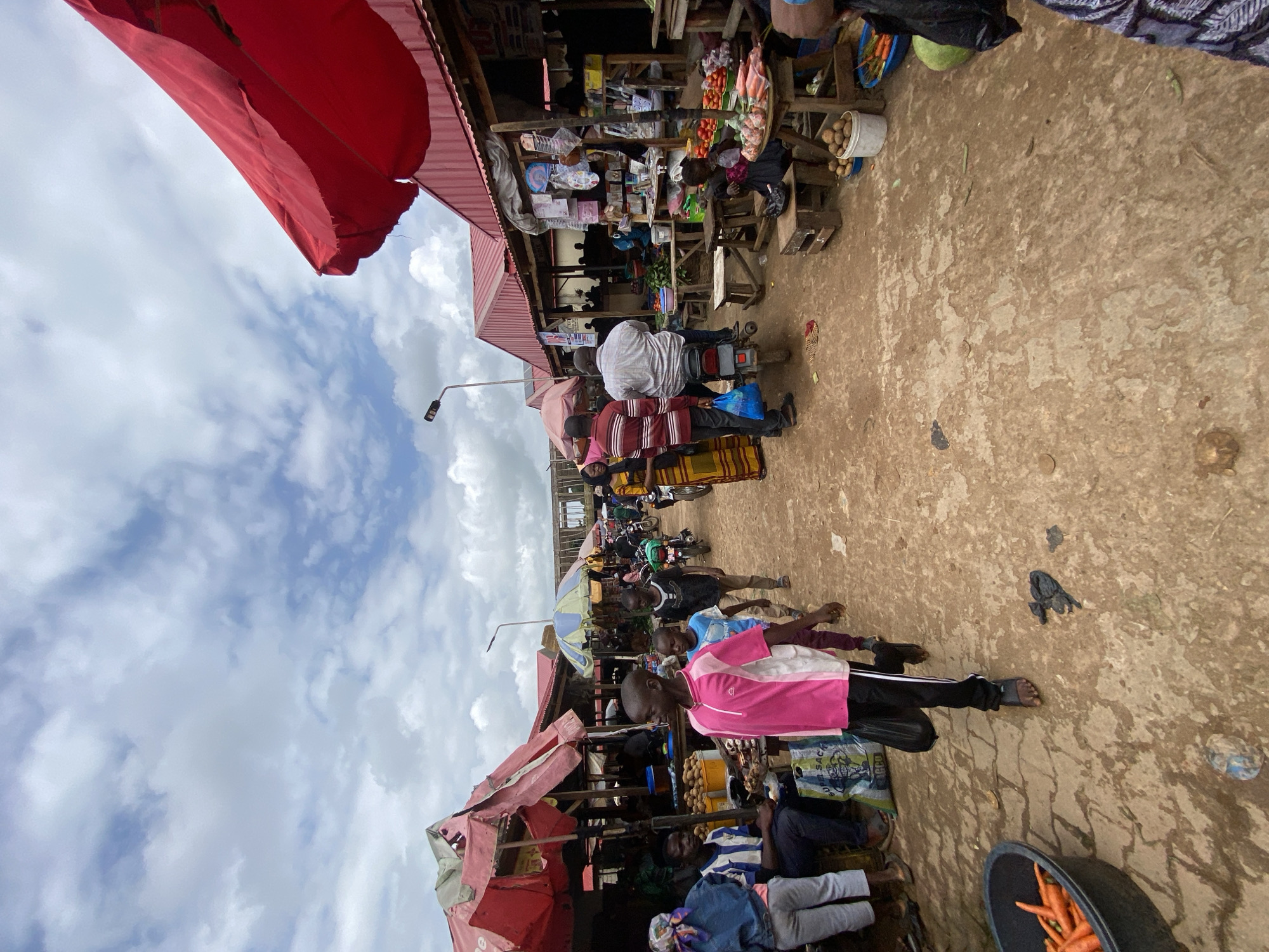 The image depicts a bustling outdoor market scene in Kafanchan B, Nigeria, under a partly cloudy sky. The market is lined with stalls covered by red and pink awnings, with corrugated metal roofs visible on some structures. The ground is a mix of dirt and what appears to be patches of wet concrete or mud.
In the foreground, a young boy wearing a pink and white t-shirt and black pants walks away from the camera, carrying a black plastic bag. To his left, a black plastic tub is filled with carrots. Further back, a group of people are gathered around stalls. One stall displays various goods, including bags of produce like tomatoes and potatoes, arranged on wooden tables and crates. Several motorcycles are parked and in use within the market.
In the middle ground, a man on a motorcycle is interacting with a person standing next to him. Other individuals are seen walking through the market, browsing stalls, and engaging in commerce. In the background, more stalls and market activity are visible, extending into the distance. The overall atmosphere is one of a lively and active marketplace.
