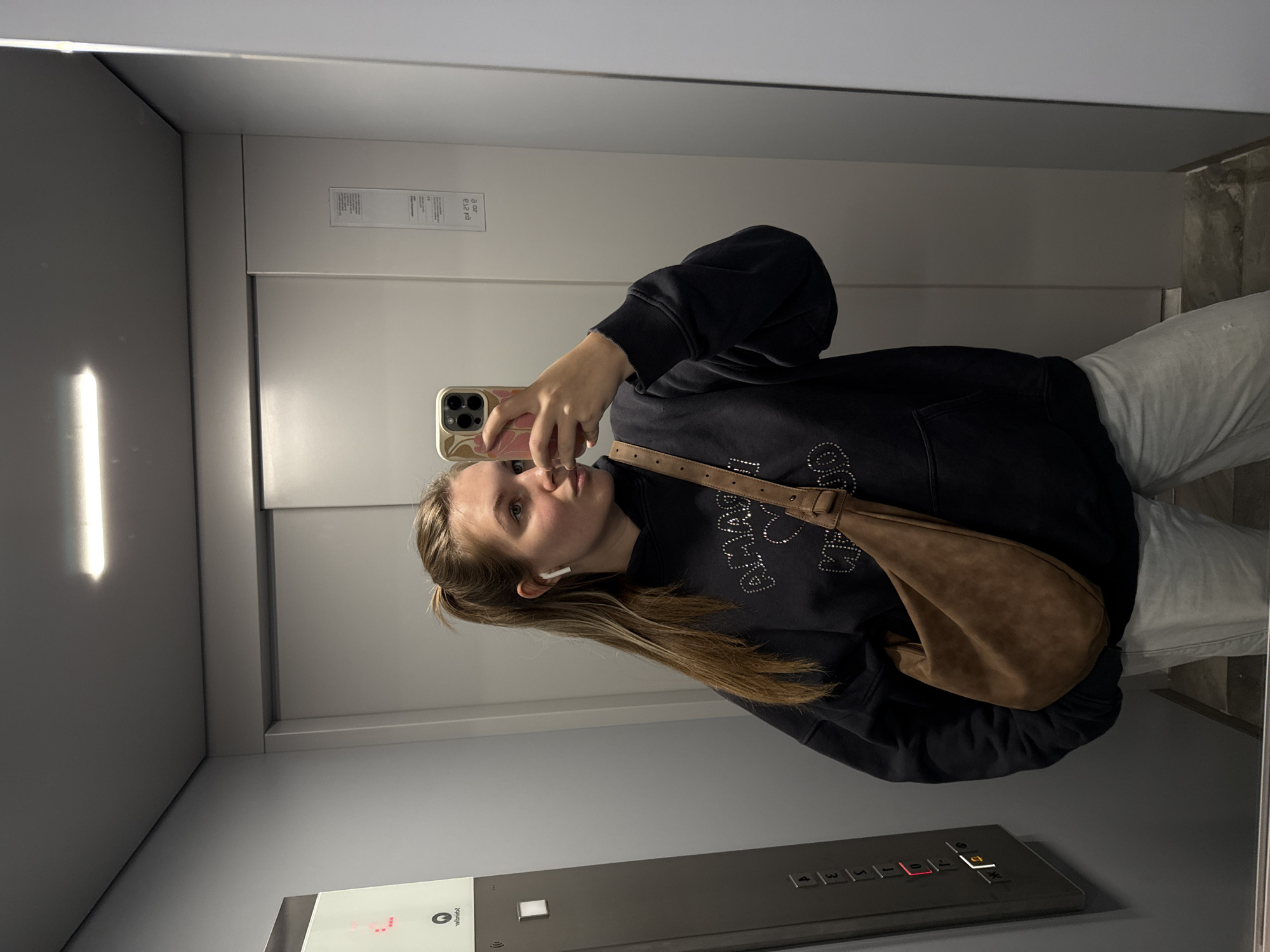 A young woman takes a mirror selfie in an elevator. She is wearing a dark hooded sweatshirt with embellishments and light-colored pants. She has a brown handbag slung across her shoulder. She is holding a smartphone in her right hand. The elevator has a button panel on the wall. Based on the location information, the elevator is in the Vozdovac Urban Municipality, Serbia. The walls of the elevator are a neutral gray color.