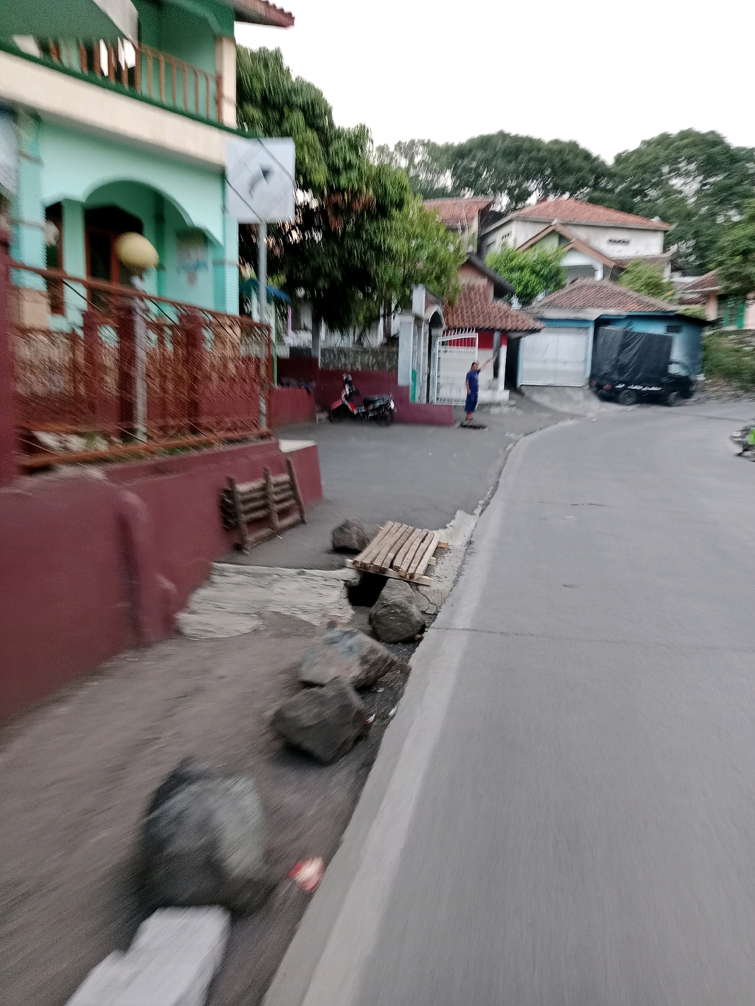 The image depicts a street scene in Cilaku, Indonesia. The foreground shows a dusty, unpaved area with several large rocks and a wooden pallet. A red retaining wall borders this area. Adjacent to the wall is a paved road that curves to the right. On the left side of the road, a light blue building with a green upper floor and a red railing is visible. A basketball hoop is mounted on the side of the building. Further down the street, several houses with tiled roofs are visible, along with trees. A person in a blue shirt and pants is standing on the sidewalk in front of a building with a closed garage door. A black truck with a tarp-covered bed is parked in front of the garage. A red motorcycle is parked on the street near the blue building. The sky is overcast.