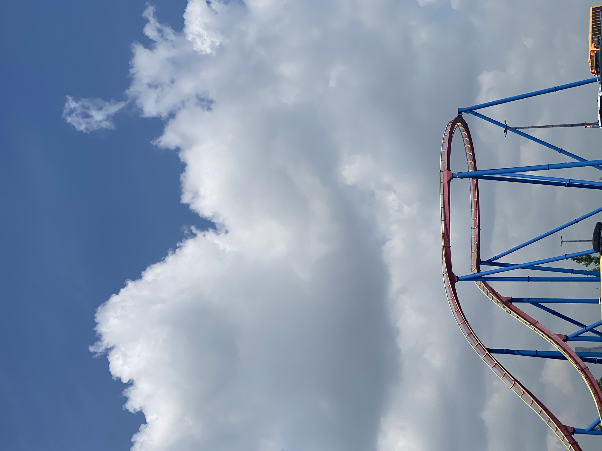 The image shows a section of a roller coaster at La Ronde amusement park in Montreal, Quebec, Canada, with a blue sky and white clouds in the background. The roller coaster track is painted red with yellow accents, and the supports are blue. The foreground shows the lower parts of the roller coaster structure, with some trees and parts of vehicles visible beneath it.