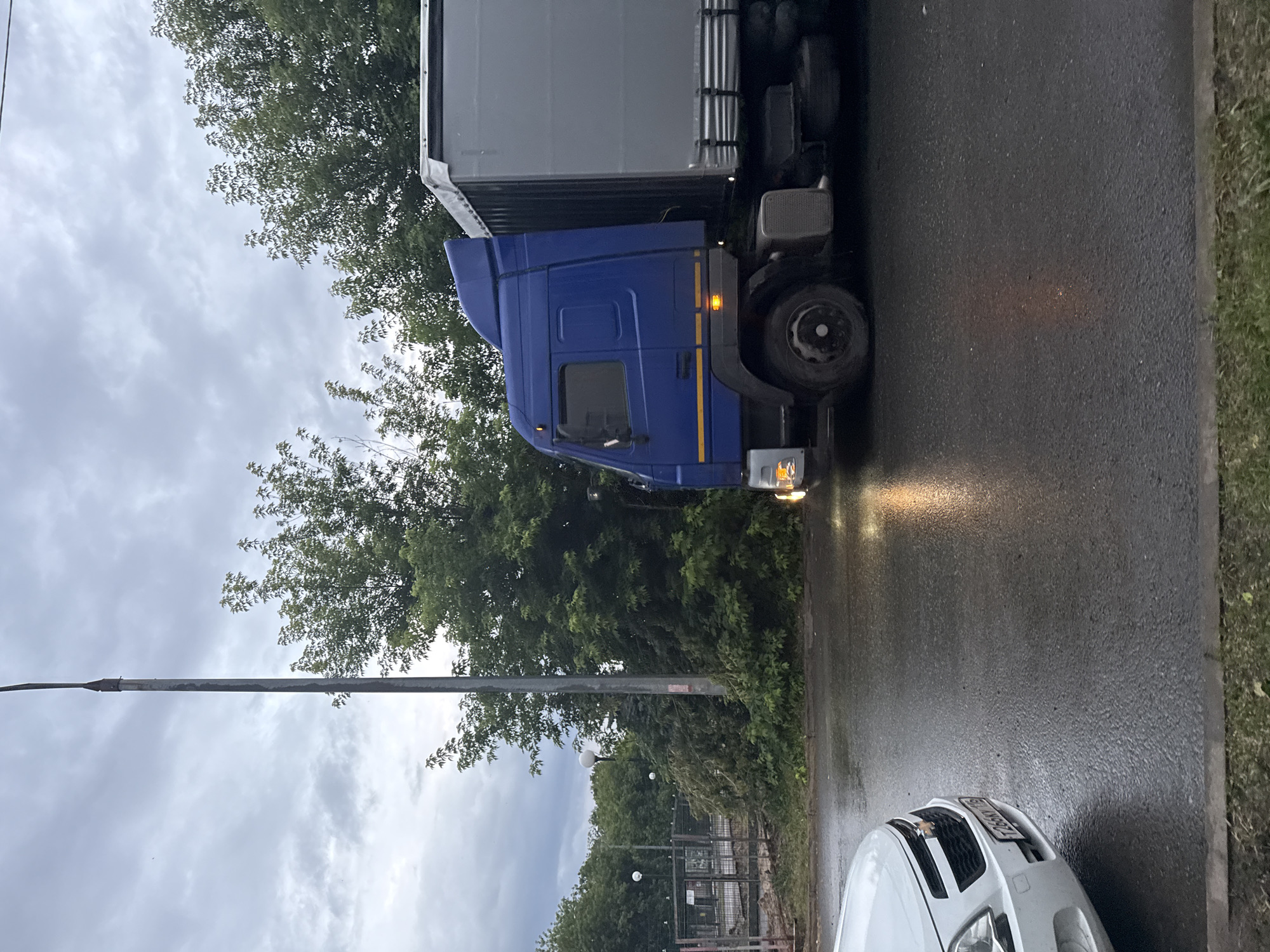 The image captures a scene on a wet, overcast day in Kazan, Russia. A blue truck with a gray trailer is parked on a paved road. The truck's headlights are illuminated, casting a warm glow on the damp asphalt. To the right of the truck, a white car is visible, its front end facing away from the viewer. The road is bordered by a grassy area. Trees with dense green foliage line the background, with a tall lamppost positioned on the left side of the frame. The sky is filled with heavy, gray clouds, suggesting recent or ongoing rainfall. A metal fence structure with lights can be seen in the distance, peeking through the trees. The overall mood of the image is somber and slightly melancholic due to the weather conditions. The license plate of the white car is partially visible and appears to be in the Russian format. No people are visible in the image.