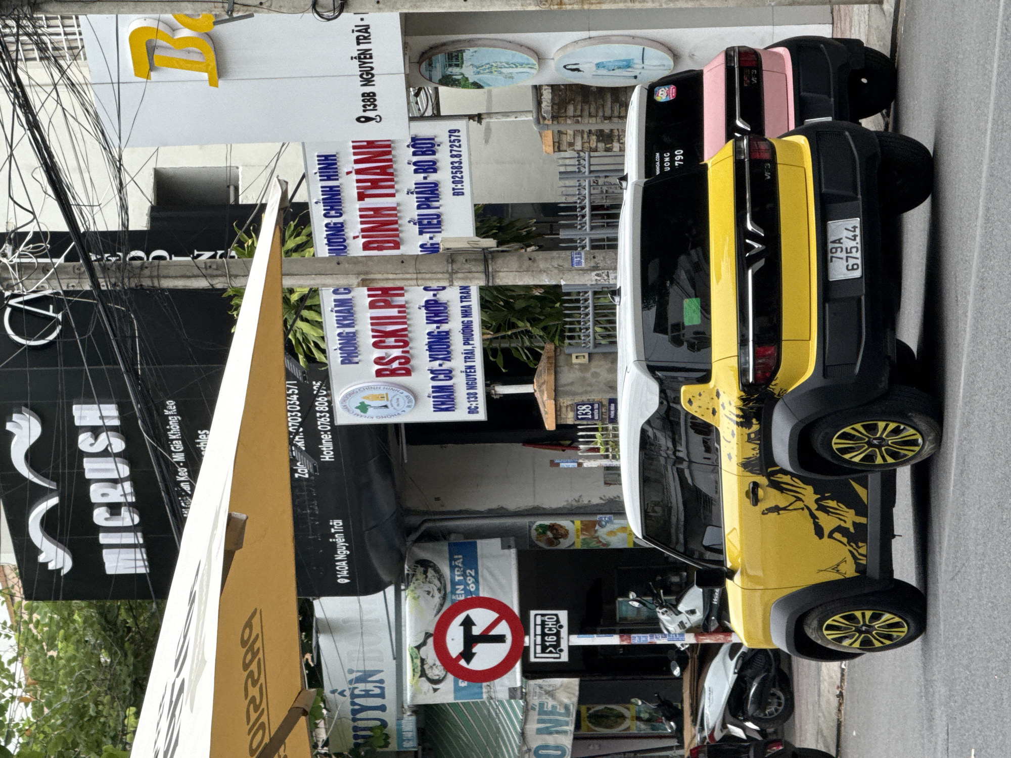 The image depicts a street scene in Nha Trang, Vietnam, focusing on two small, modern cars parked on the roadside. The car in the foreground is bright yellow with black accents and a striking black graphic depicting a mountainous landscape with a silhouetted figure hiking. The car behind it is pink. Both vehicles have their rear ends facing the viewer, with visible license plates and taillights.
The background features a sidewalk with several shops and businesses. Prominent signage includes "MICRUSH," "CA," and a yellow "B." One shop has a sign that appears to be a medical clinic with Vietnamese text, including "PHÒNG KHÁM C" and "ĐÌNH THÀNH." Another sign displays a traffic symbol with a red circle and a line through a left-turn arrow and a straight arrow, indicating a restriction on turns, along with a bus symbol and the text ">16 CHỖ" (over 16 seats). Streetlights, utility poles with a dense network of wires, and trees are also visible. Several motorcycles are parked on the sidewalk. The overall impression is a busy urban environment.