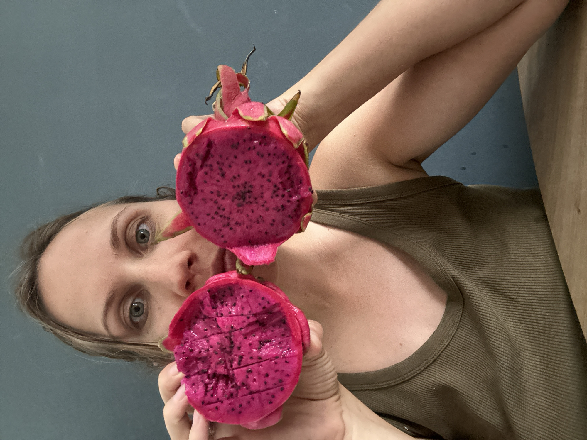 A person is holding two halves of a vibrant pink dragon fruit in front of their face. Their eyes, which are wide and looking at the camera, are visible between the two halves of the fruit. The fruit is cut in half, revealing its bright pink flesh speckled with small black seeds. The person is wearing a brown tank top. The background is a plain, dark blue wall. The lighting suggests it is daytime, and there are no immediate cues about the weather or specific location other than the provided context of Fethiye, Turkey. The overall emotion conveyed is one of playful curiosity. There is no visible text in the image.