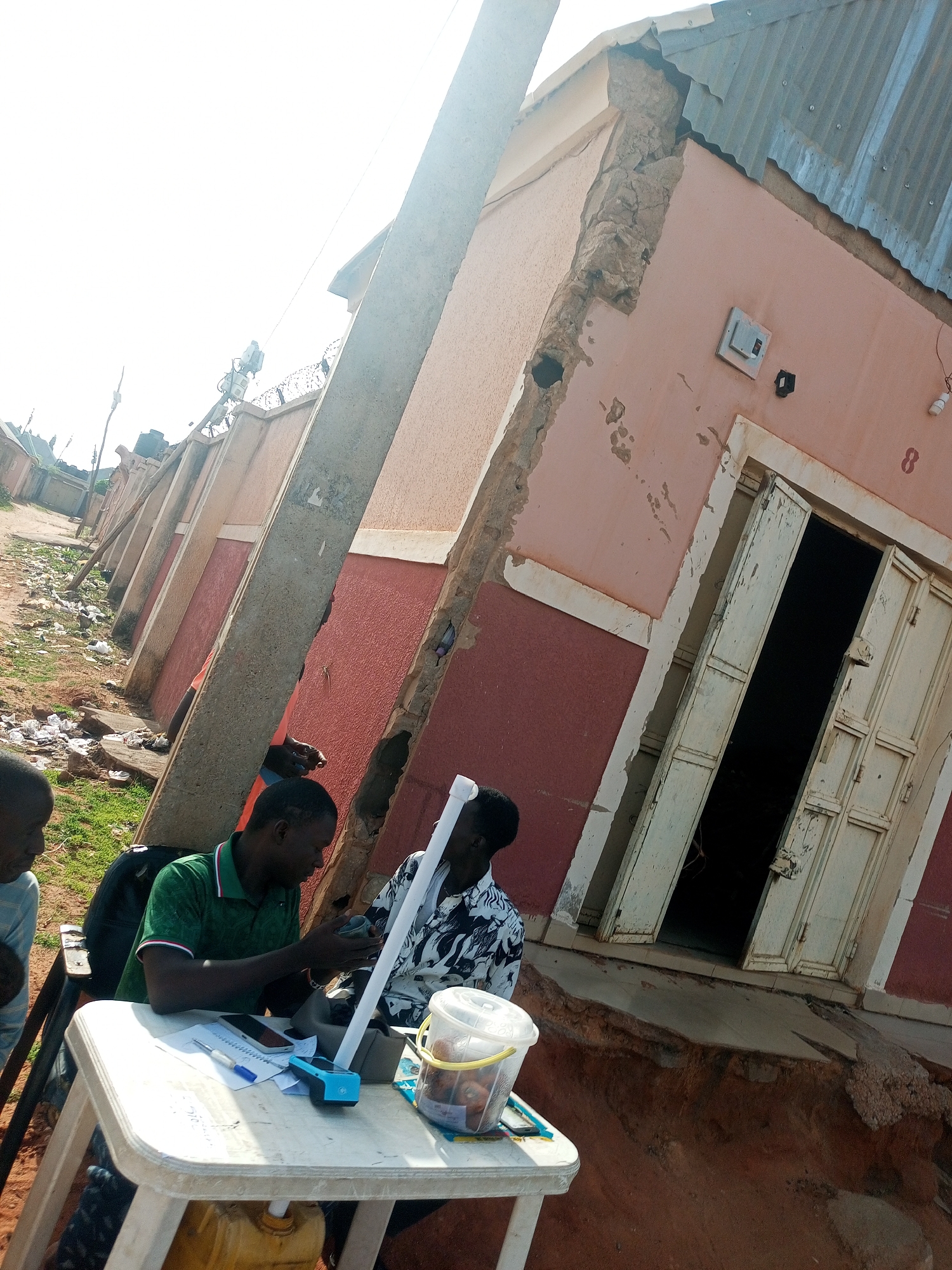 The image shows an outdoor scene in Gombe, Nigeria. Two men are seated at a white plastic table next to a street or roadside. One man wears a green shirt, and the other wears a patterned black and white shirt. On the table, there are papers, pens, a blue device, a cell phone, and a translucent plastic bucket with contents. A yellow jerrycan is visible under the table.
Behind them is a row of pink and red buildings or structures. A concrete utility pole stands at an angle. The building closest to the table has an open, weathered door, and the number "8" painted on the wall. The building's construction appears to be in disrepair, with exposed materials. The ground in front of the building is eroded. A fence with barbed wire runs along the top of the buildings. Other buildings can be seen further down the street. The overall impression is one of a community facing environmental or structural challenges.