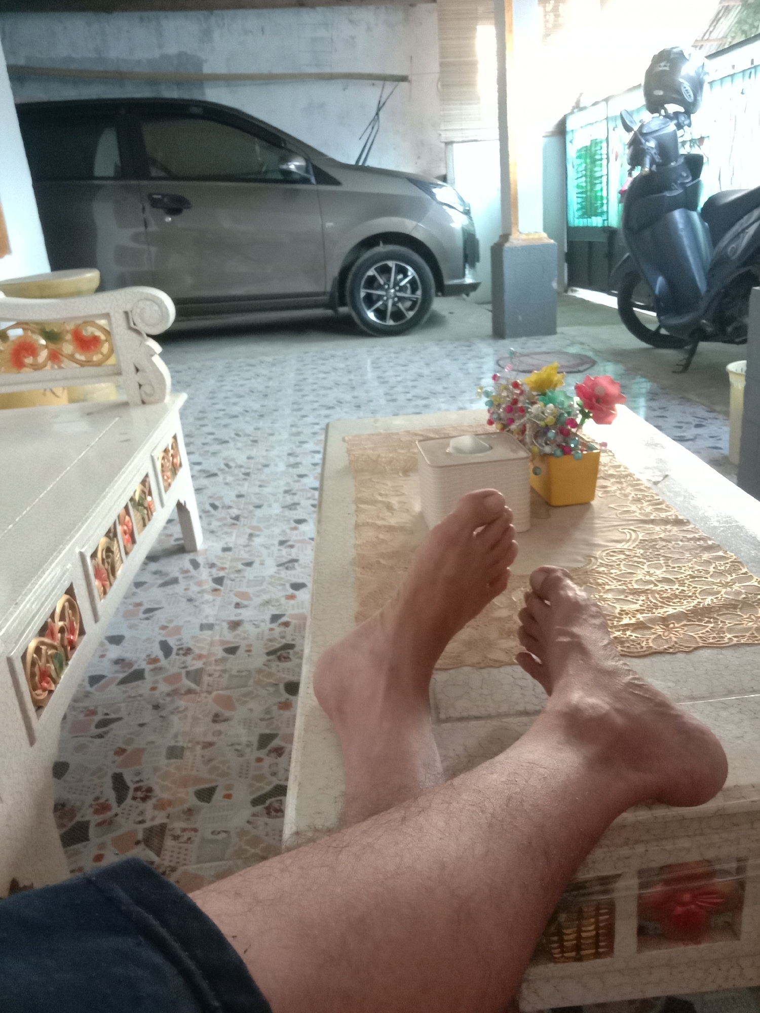 In Kwandang, Indonesia, a person's bare feet and lower legs are visible, resting on a tiled floor. The person is seated in what appears to be a covered outdoor area, possibly a garage or patio. In the background, a light-colored car is parked, and to the right, a black motorcycle with a helmet on its handlebars is visible. A small table in the foreground holds a tissue box and a decorative vase with colorful artificial flowers. A white ornate bench is positioned to the left of the frame. The floor is covered in terrazzo tiles with a multi-colored speckled pattern.