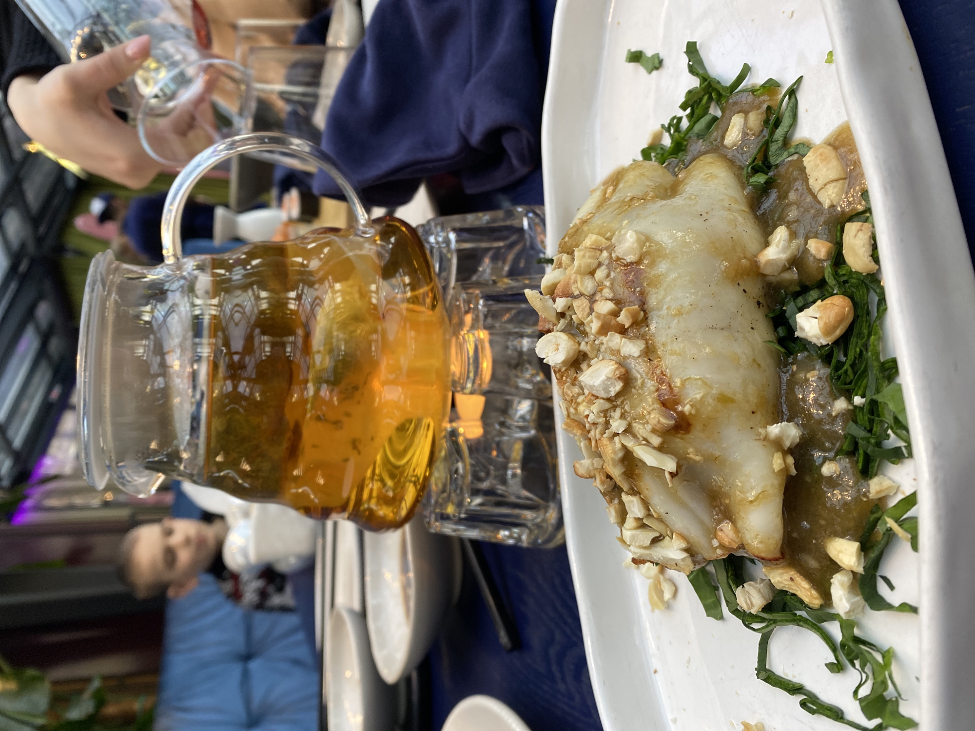 Here is a detailed description of the image:
**Scene:** The image shows a table setting, likely in a restaurant, with a plate of food and beverages.
**Food:** A white oblong plate holds a dish consisting of a cooked piece of what appears to be a protein, possibly fish or chicken, topped with chopped nuts and sitting on a bed of green leafy vegetables and a sauce.
**Beverages:** Two clear glass pitchers or mugs, one filled with a golden liquid that could be beer or iced tea. There is also a glass in the background.
**People:** A blurred image of a child is visible in the background. A hand is holding a glass.
**Objects:** Additional plates and cutlery are partially visible on the table.
**Environment:** The setting appears to be indoors, possibly in a restaurant. The background features blue seating and some plants, suggesting a casual or semi-formal atmosphere.
**Location Information:** The image does not provide enough information to determine if the location is in the city of Yekaterinburg, Russia. Additional information would be needed to confirm this.