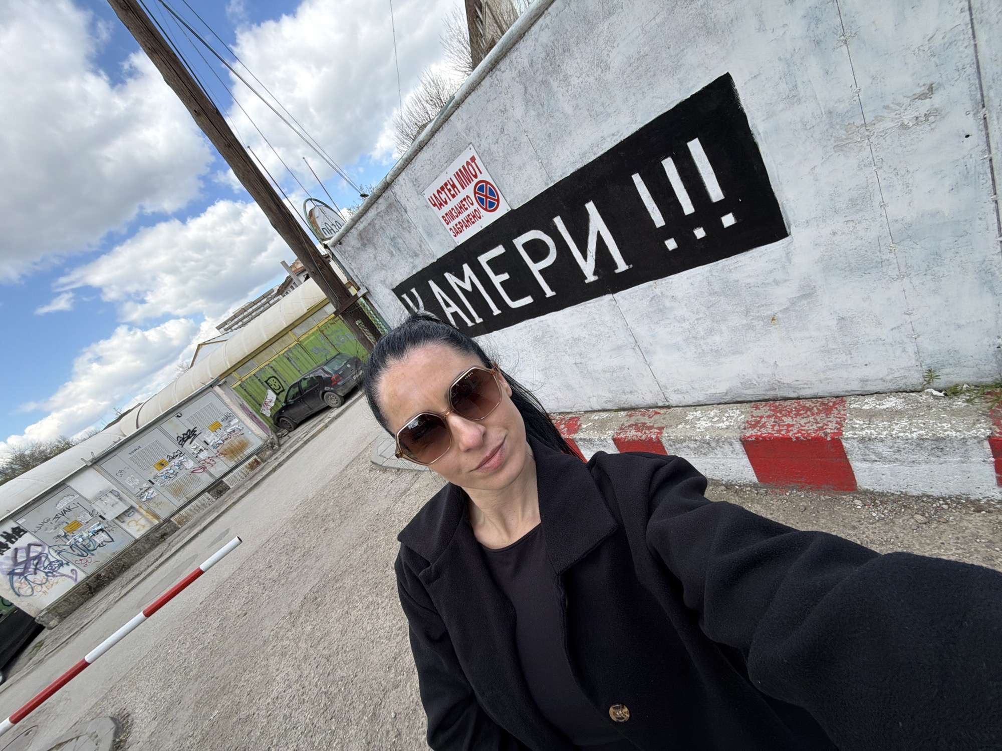 The image depicts a woman wearing sunglasses and a black jacket, taking a selfie outdoors. Behind her is a weathered white wall with a black rectangle containing the word "КАМЕРИ !!!" (Cameras !!!) written in Bulgarian. Above this, there is a sign that reads "ЧАСТЕН ИМОТ! ВЛИЗАНЕТО ЗАБРАНЕНО!" (Private Property! No Entry!). The wall is topped with a painted red and white barrier.
In the background, there are industrial buildings, some covered in graffiti, and a parked car. Utility poles and wires are visible in the sky above.
Based on the Bulgarian language signage, the location is in Bulgaria. Considering that the individual taking the photo has also previously taken photos around Pleven, Bulgaria, I would estimate that this is also the location of the current photograph. The specific street is likely to be on the outskirts of the city center, in an industrial or commercial area.