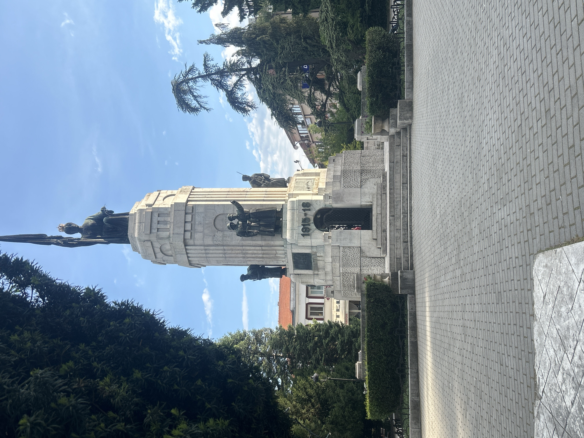 The image shows the Monument to the Asenevtsi in Veliko Tarnovo, Bulgaria. It is a large, white stone monument featuring bronze sculptures of several figures, including soldiers and a winged figure. The monument stands on a tiered platform with steps leading up to it. A paved walkway borders the monument, and there are bushes and trees surrounding the structure. In the background, buildings are visible under a blue sky with scattered clouds.