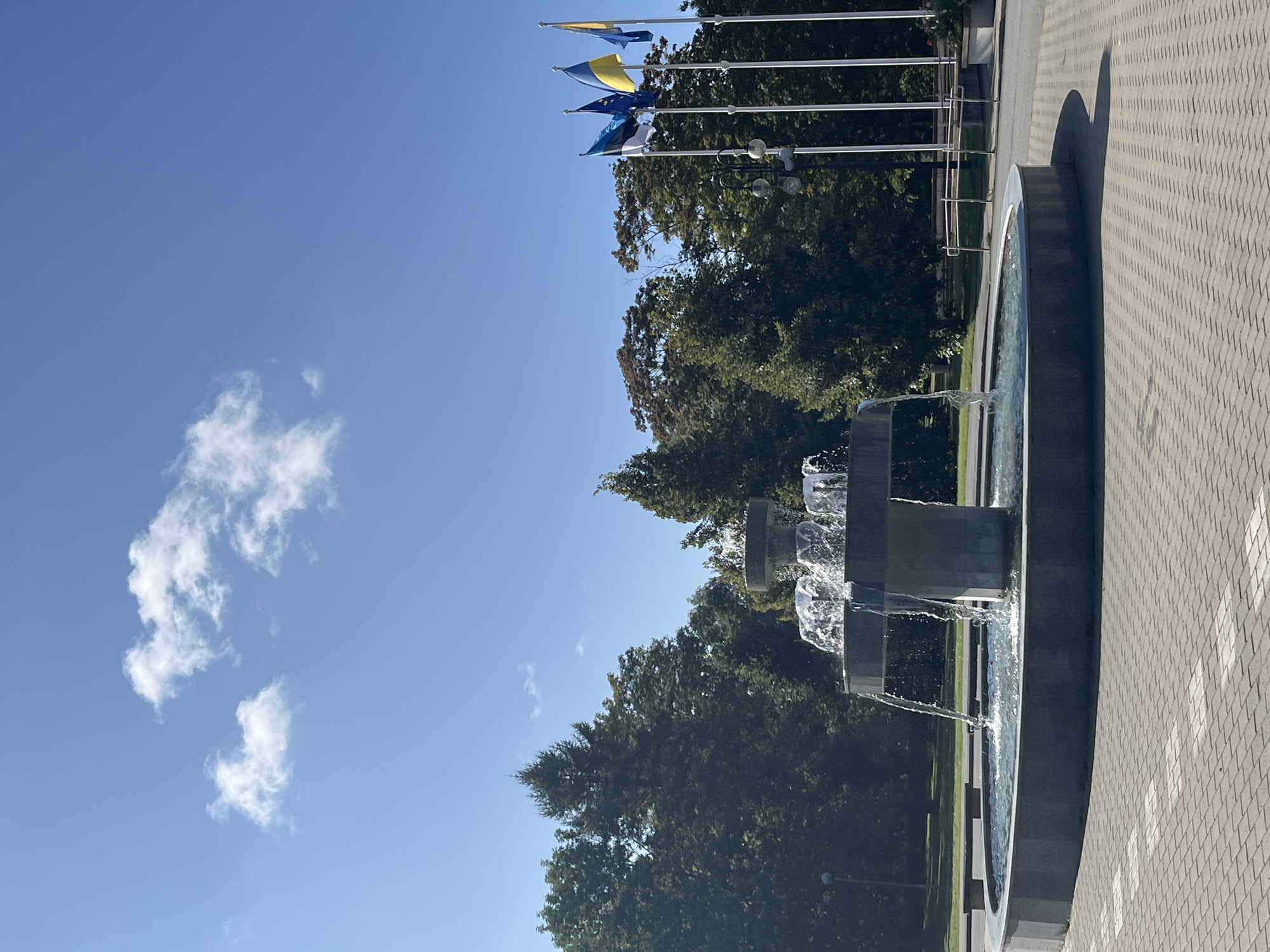 The image shows a public square with a tiered fountain in the foreground. The fountain is made of dark gray stone and has water cascading from its upper levels into the lower basin. Surrounding the fountain is a paved area with a pattern of light and dark gray interlocking bricks. In the background, there are tall trees and a clear blue sky with a few scattered white clouds. To the right of the fountain, there are several flagpoles flying flags, including what appears to be the flag of Estonia, the European Union flag, and the flag of Ukraine. The scene suggests a sunny day in an outdoor public space. The location is the city of Kohtla-Järve linn, Estonia. There are no people visible in the image.