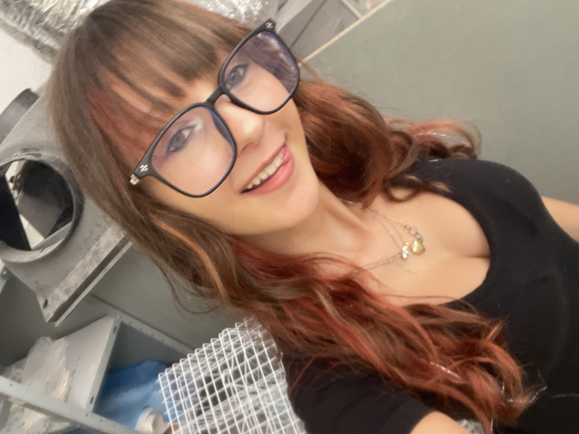 In an indoor setting, possibly a storage or workshop area, a young woman with auburn hair, streaked with pink and blonde highlights, takes a selfie. She is wearing a black V-neck top and several necklaces, including one with a heart-shaped pendant. She has on black-rimmed glasses and is smiling, with her tongue slightly visible between her teeth. In the background, metal shelving units are visible, one containing a blue plastic bag. A white wire rack is also present. The overall lighting is somewhat dim, with the light source appearing to be from above and to the right. The location is identified as Raanana, Israel, though no specific landmarks or indicators of the city are apparent in the image itself.