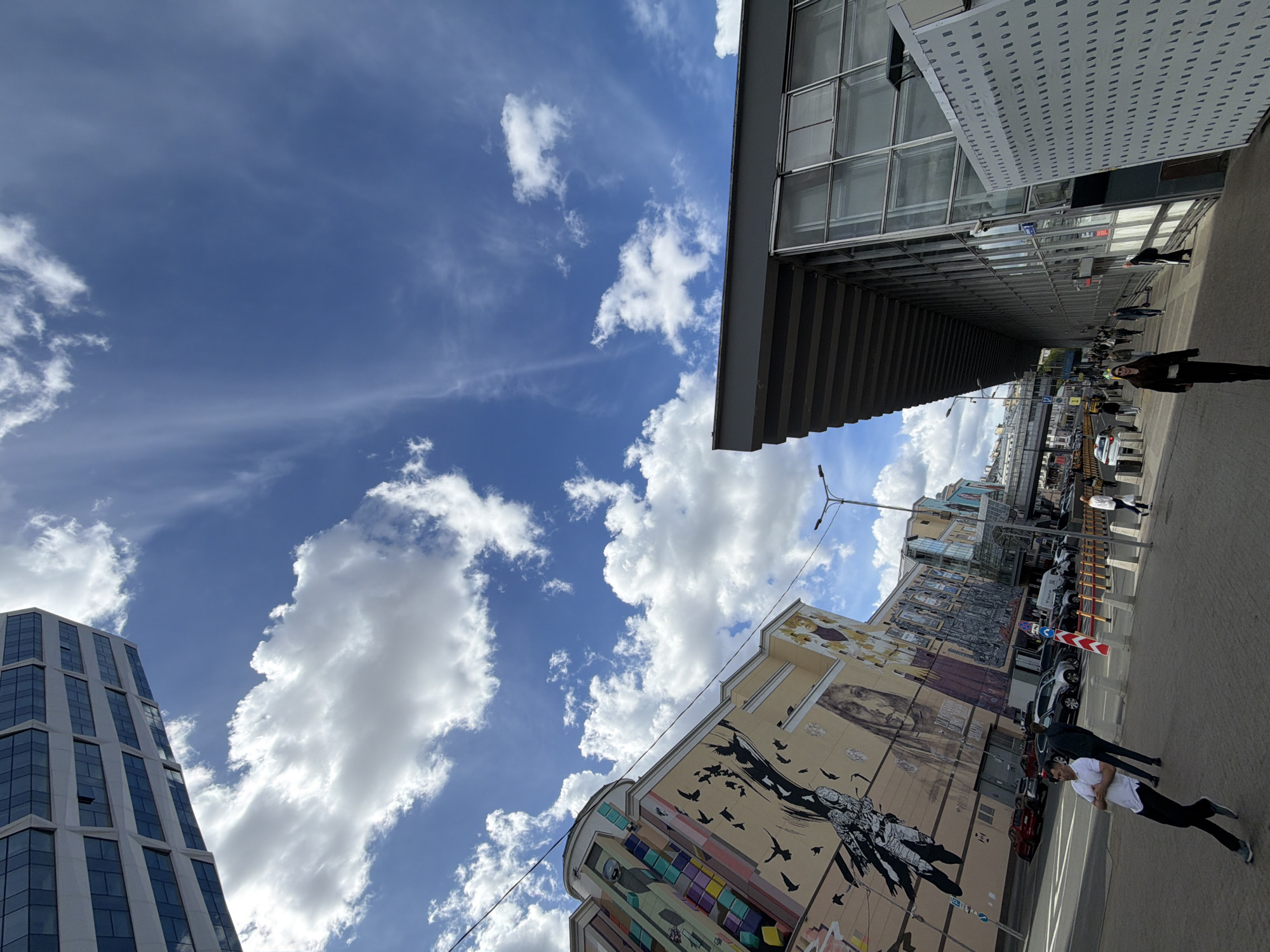 The image captures a street scene in an urban environment. Buildings frame the sky, and one building features a large mural on its side. People walk along the sidewalk next to a street with parked cars and passing vehicles. Streetlights and traffic signs are visible among the buildings. The sky is blue with scattered clouds.