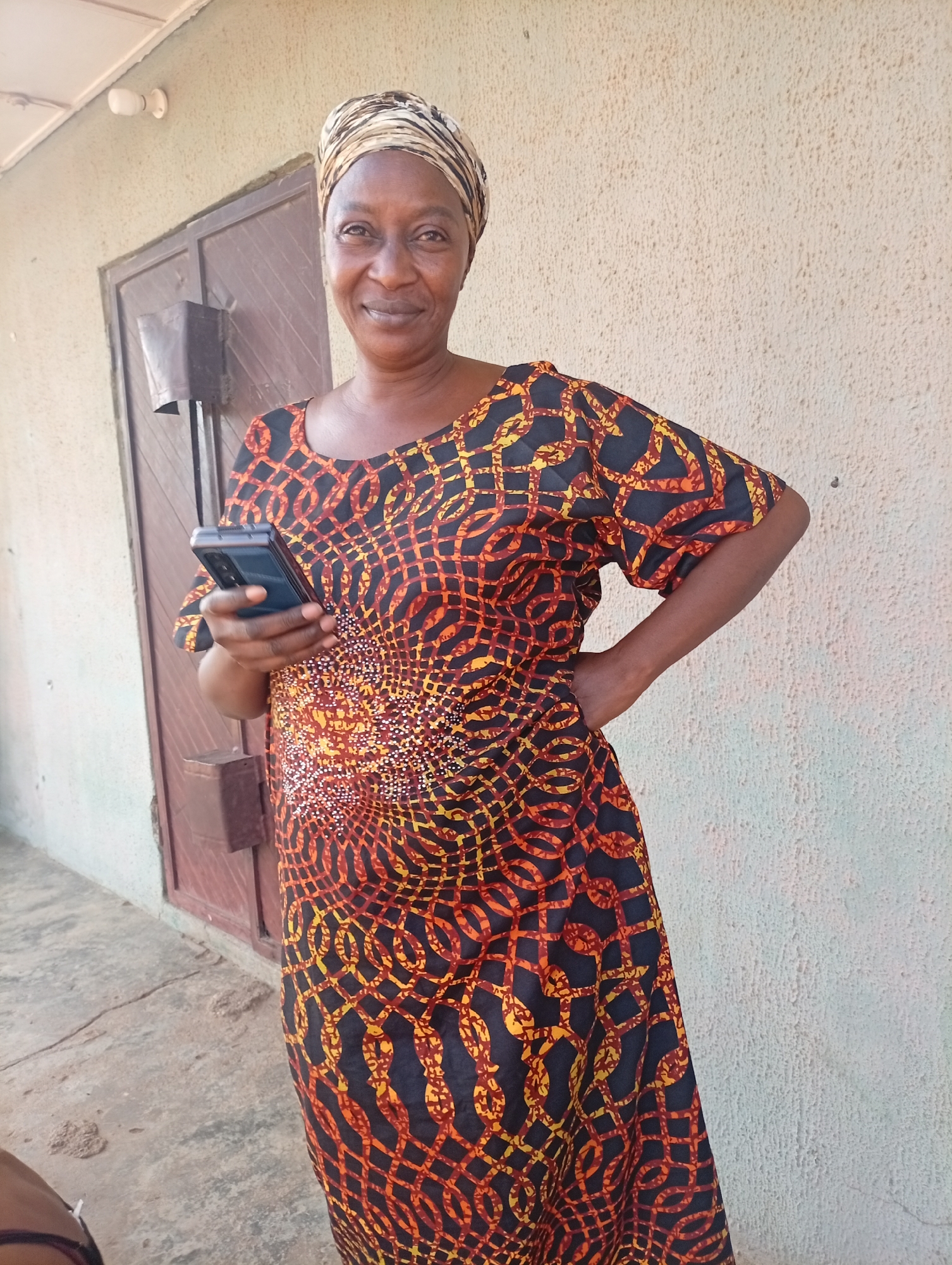 A middle-aged African woman with dark skin, a light brown headwrap, and a colorful patterned dress stands in front of a textured wall with a doorway to her left. She is holding a black smartphone in her right hand and has her left hand on her hip. The woman is smiling and looking directly at the camera. The environment appears to be outdoors, possibly in a residential area of Sabon Tasha, Nigeria, given the architectural style and the rough ground surface. A lightbulb is visible on the wall above the doorway.
