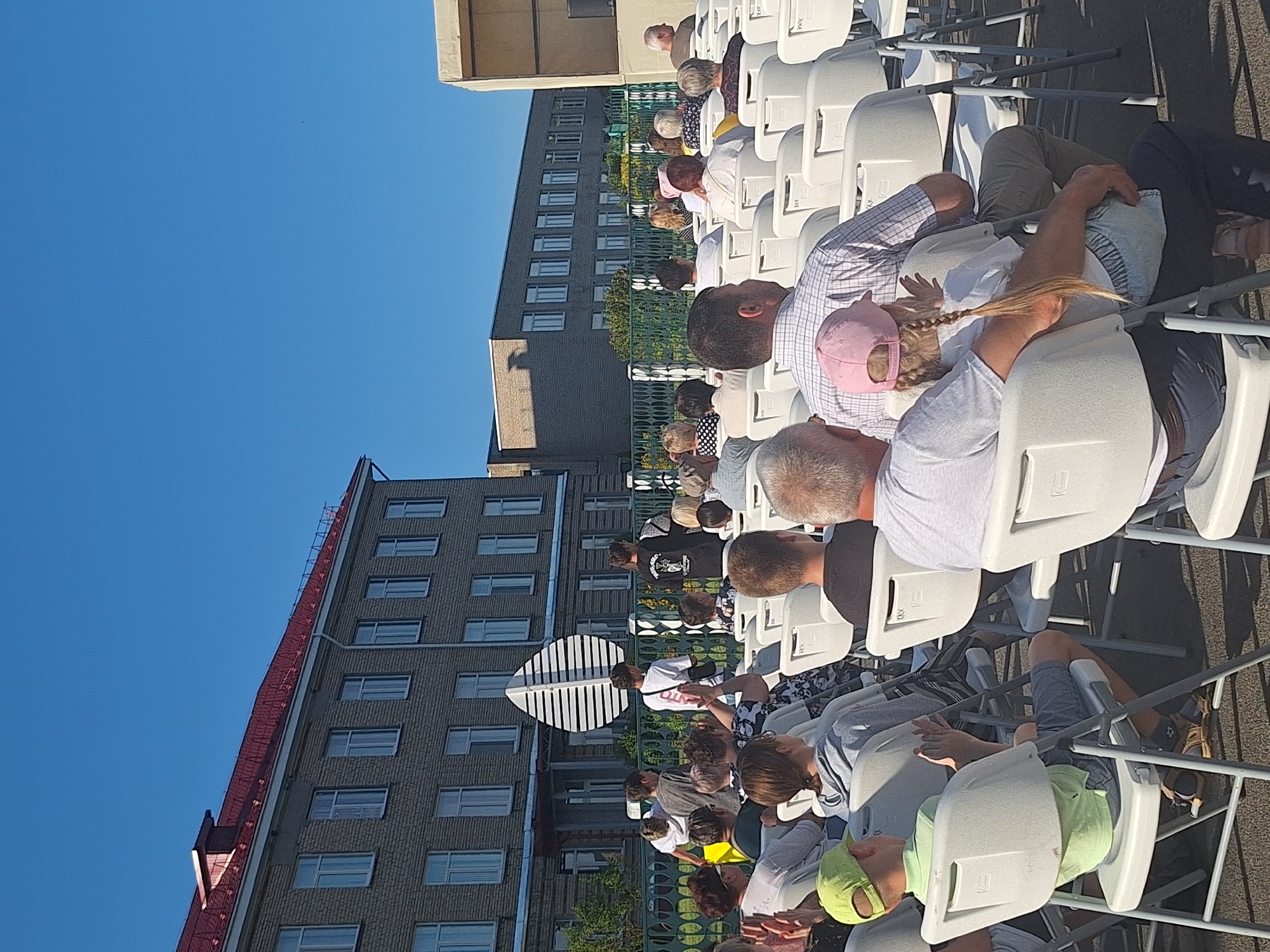A group of people are seated in white plastic chairs outdoors, facing a stage area that is partially visible on the right side of the frame. The setting appears to be a school or community event, with a large brick building with a red roof in the background. The sky is clear and blue, suggesting it is a sunny day. Some individuals are looking towards the stage, while others are interacting with each other. The overall atmosphere seems relaxed and social.