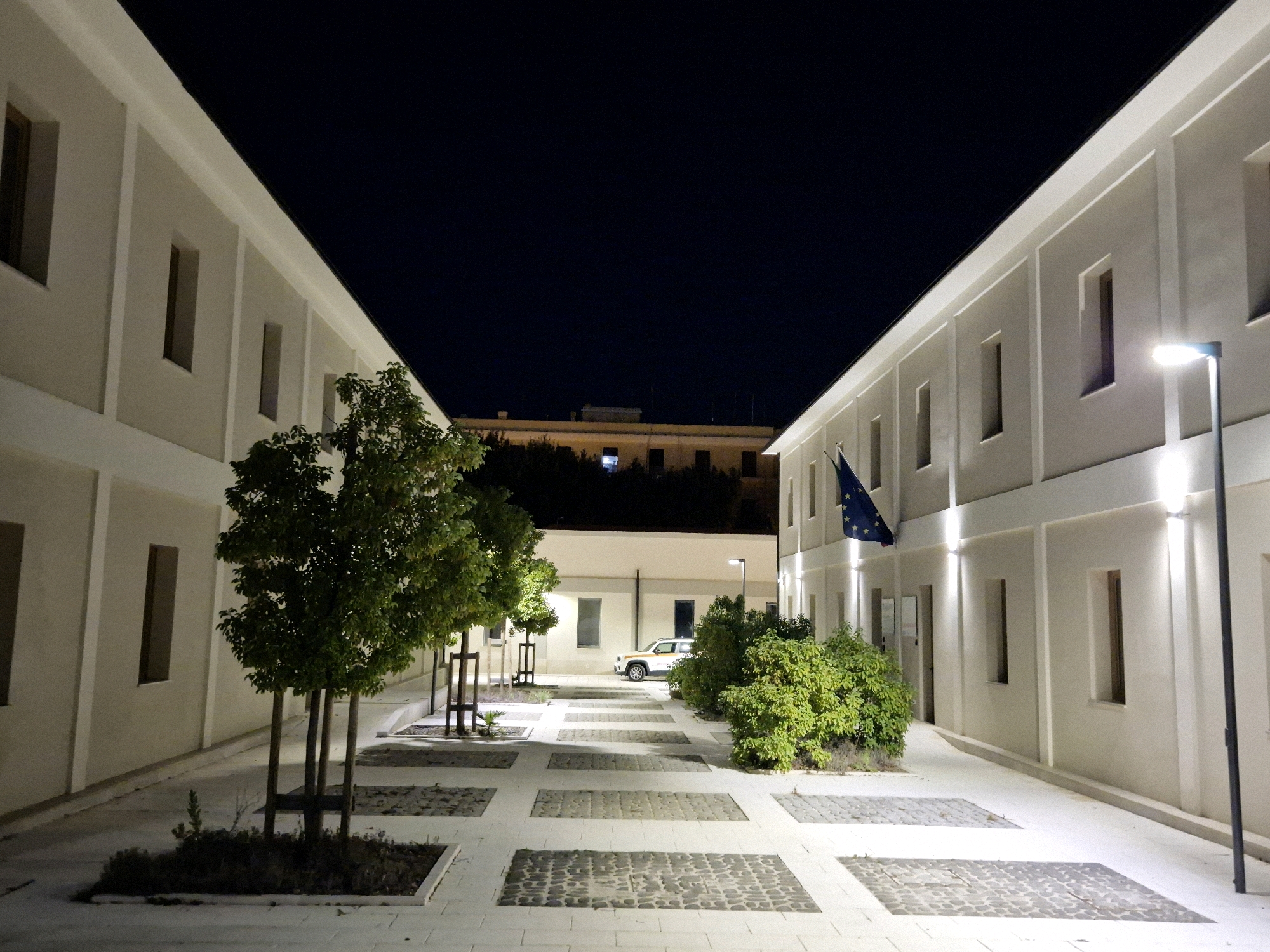 The image depicts a modern courtyard at night, likely part of an institutional building, in the city of Taranto, Italy. The courtyard is flanked by two long, pale buildings with a minimalist architectural style, featuring numerous vertical rectangular windows. The ground is paved with white tiles interspersed with rectangular sections of cobblestones.
On the left side, several young trees with green foliage are planted in square garden beds. On the right side, a dense green bush is visible. The background shows a darker building with illuminated windows and the silhouette of trees. A white car is parked in the middle ground. A flag, identified as the European Union flag, is prominently displayed on the right-hand building, fluttering in the breeze. Exterior lighting fixtures illuminate the facade of the buildings and parts of the courtyard, creating a stark contrast with the dark night sky. The scene conveys a sense of order and modern design.