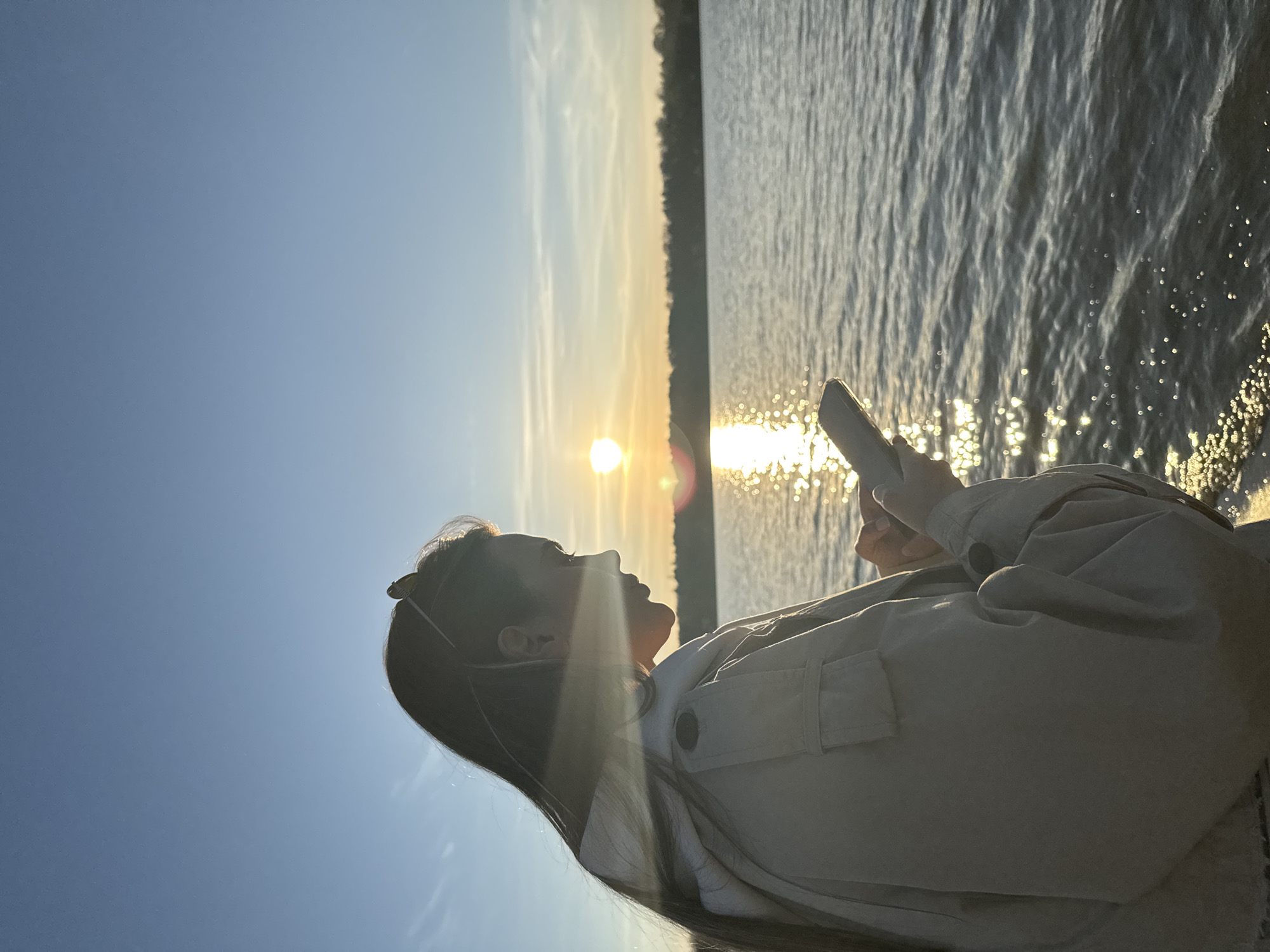 A woman is silhouetted against a sunset over water. She is holding a mobile phone, looking towards the sun. The scene includes the sky, water reflecting sunlight, and a distant shoreline. The location is Arciechów, Poland.