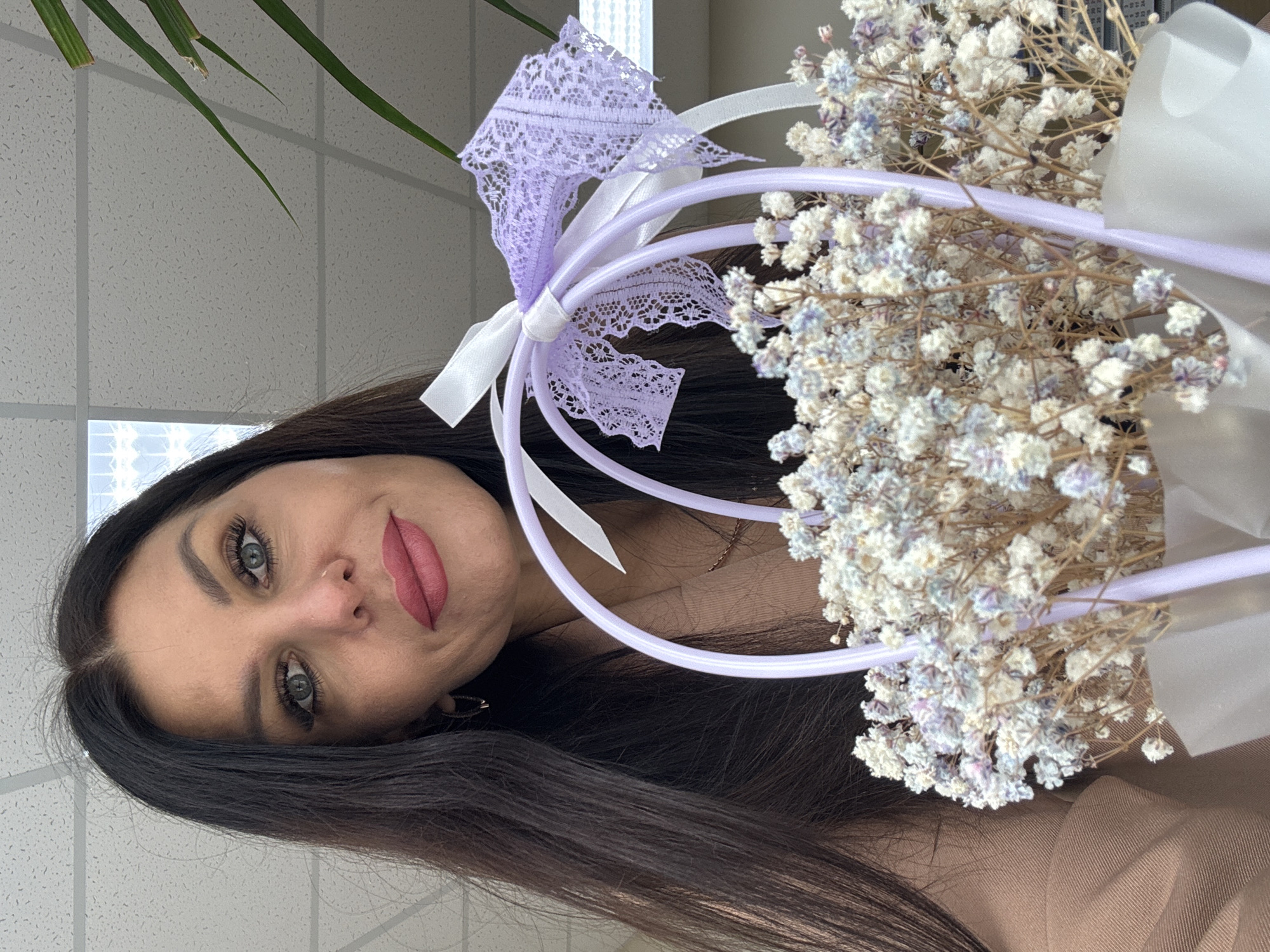 The image shows a woman with long, dark hair, fair skin, and a neutral facial expression, holding a bouquet of small white flowers with lavender accents, wrapped with white ribbons and lace. The bouquet is supported by a lavender-colored frame. The background consists of a white ceiling with a grid pattern, suggesting an indoor setting like an office or institutional building. A partial view of a green plant is visible in the upper left corner. While the location cannot be determined based on the image, the prompt provides information that the picture was taken in the city of Chaikovsky, Russia.