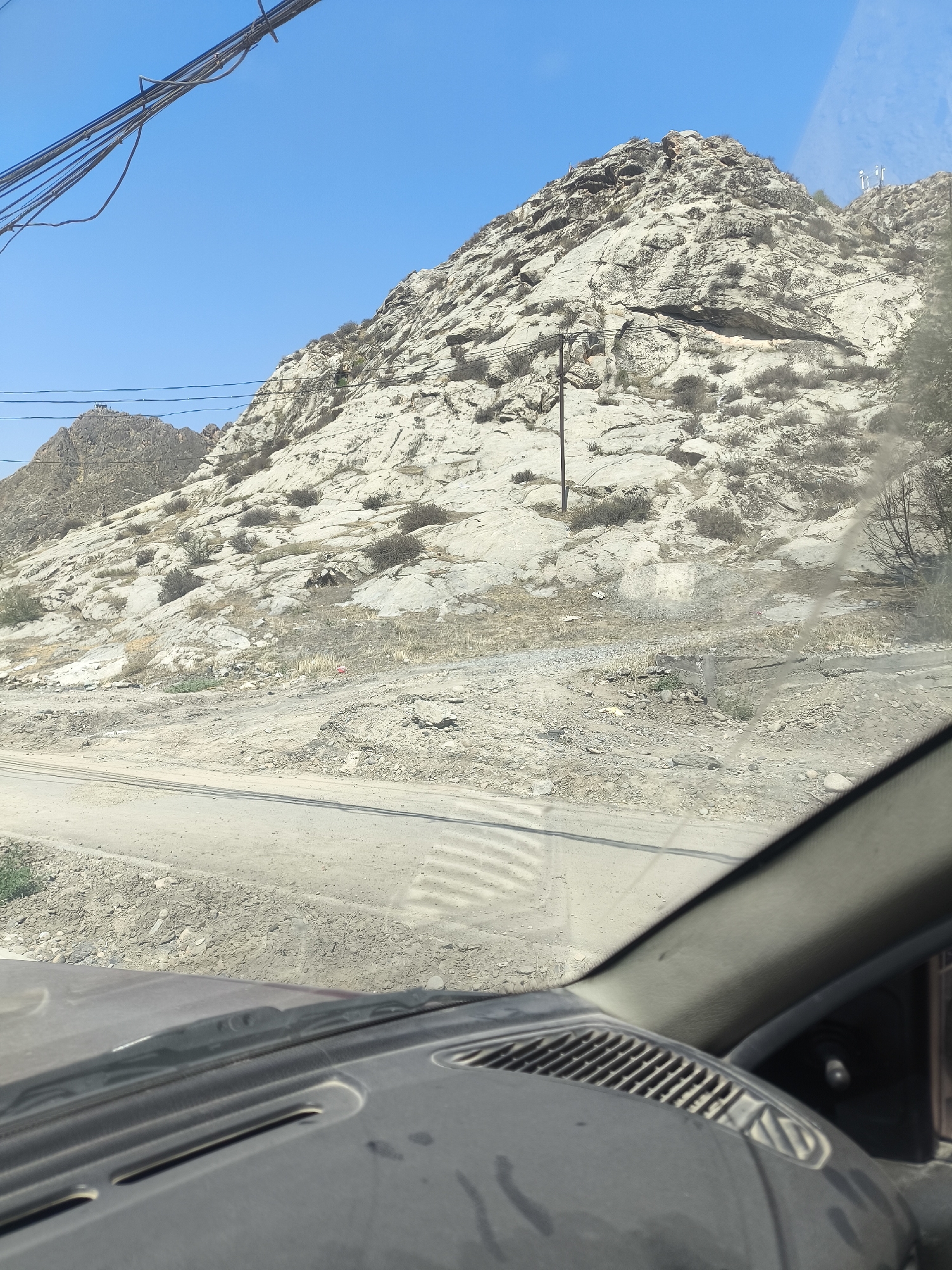 The image, taken from inside a vehicle, shows a rocky, mountainous landscape under a clear blue sky. Power lines are visible in the upper left corner. The terrain is arid with sparse vegetation, and a dirt road runs through the foreground. A single utility pole stands on the mountainside. The interior of the car, including the dashboard and windshield, is visible, suggesting the photograph was taken while driving. There are no people or animals present in the scene. The location is not specified.