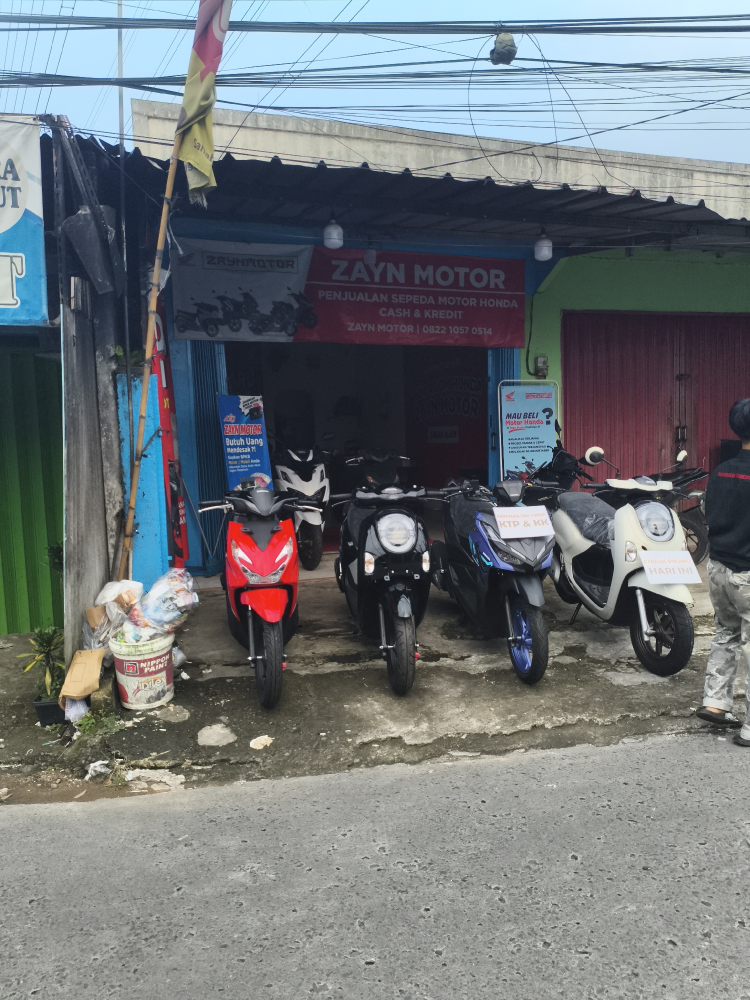The image shows the front of a motorcycle dealership named "Zayn Motor" in Depok, Indonesia. A red banner above the shop's entrance advertises "Penjualan Sepeda Motor Honda Cash & Kredit" (Honda motorcycles for sale, cash and credit). Several motorcycles are parked in front of the store: a red scooter, a black scooter, a blue and black scooter, and a white scooter. A sign reading "MAU BELI Motor Honda" (Want to buy a Honda motorcycle?) is on display. A bucket of Nippon Paint is located near the storefront's left side. Power lines are visible above the building. A partially obscured person is standing to the right of the scooters, near the sidewalk.