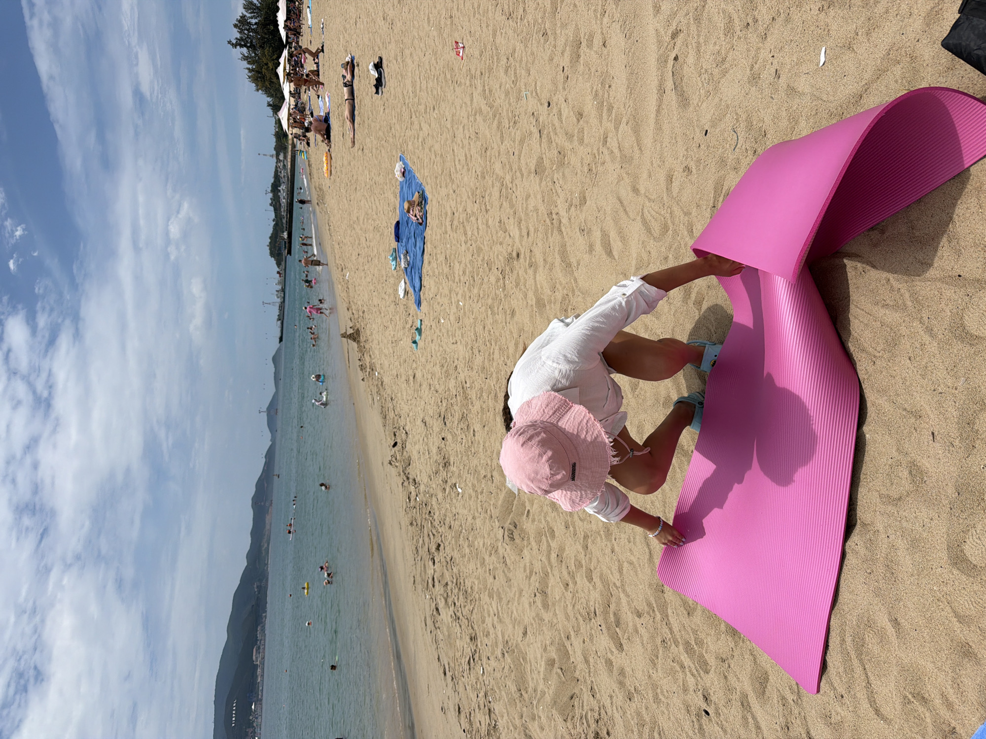 In Nha Trang, Vietnam, a person wearing a pink hat and a white shirt is on a sandy beach, crouched over a bright pink yoga mat. The mat is partially unrolled and appears textured, with ripples running across its surface. The person is reaching down towards the mat. The beach is populated with other individuals, some in the water and others sunbathing on towels. The sea is calm with distant boats and a mountainous coastline under a partly cloudy sky. The overall scene captures a leisurely day at the beach.