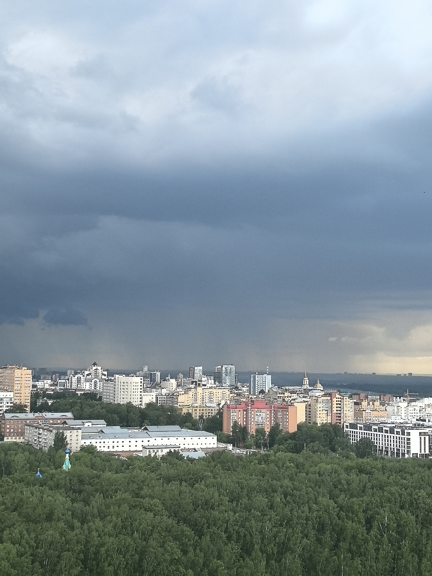 The image presents a high-vantage wide shot of a city and a dense forest under a dramatic, stormy sky. In the immediate foreground, a vast expanse of lush, green deciduous trees dominates, suggesting a large park or natural area. Within this forest, near the left, two small, light blue, conical structures with golden tops, possibly parts of a church or gazebo, peek through the canopy.
Beyond the forest, the cityscape unfolds, featuring numerous multi-story buildings of varying architectural styles and colors. There are older residential blocks with muted facades (brown, yellow, white) alongside taller, more modern structures. Several distinctive spires and domes rise above the general skyline in the mid-distance. The horizon line, visible beyond the buildings, shows a distant landscape or body of water under a lighter, hazy sky.
The most striking feature is the sky, which is heavily laden with dark, grey storm clouds that cover most of the upper frame. In the distance, rain is visibly falling from these ominous clouds over the city and beyond, appearing as faint vertical streaks. The overall lighting is dim, indicative of a cloudy or stormy day, likely late afternoon or early evening given the muted light. The scene captures the moment of a significant weather event gathering over an urban environment. No people are discernible, and no text is visible. The urban architecture and natural setting are consistent with a large city like Perm, Russia.