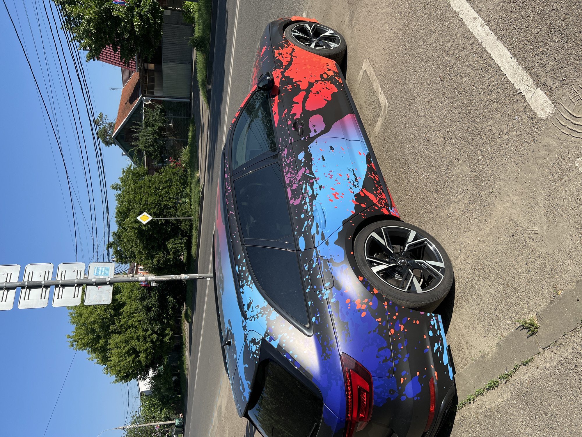 A custom-wrapped Audi RS4 Avant (B9 generation) is prominently displayed, parked on a paved surface adjacent to a road in the city of Krasnodar, Russia. The vehicle features a matte black base wrap, intricately covered with a gradient of vibrant paint splatters: indigo and blue dominate the rear, transitioning through purple to vivid red and pink towards the front. The car is equipped with multi-spoke alloy wheels, displaying the Audi rings logo.
The immediate foreground consists of a coarse asphalt and gravel surface with visible cracks and sparse grass growth. A white solid line marks the edge of the parking area, and two square utility covers are embedded in the pavement.
To the left of the vehicle, a grey metal utility pole stands, featuring five stacked, blank, rectangular white signs and one smaller, partially obscured sign with faint blue and red text. Further along the road, a yellow diamond-shaped road sign, indicating a priority road, is visible.
The background reveals a residential street scene under a clear blue sky. A two-lane road with a dashed white center line runs parallel to the parked car. Beyond the road, dense green foliage from trees and bushes partially conceals several houses, some with distinct red-tiled roofs and one with green siding. Numerous black power lines are strung across the upper portion of the frame, traversing the sky. No individuals are visible in the scene.