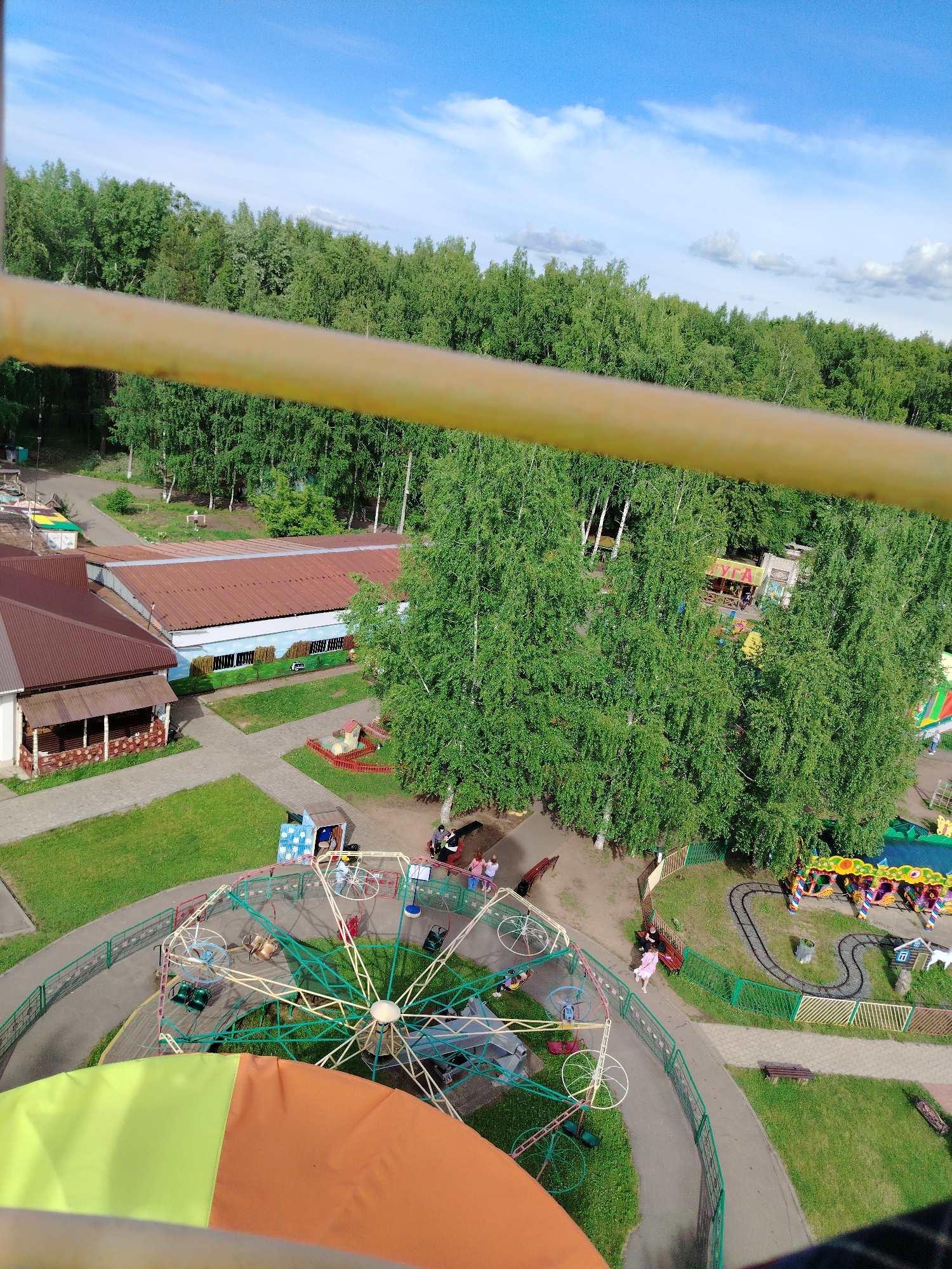 This high-angle image captures an aerial view of an amusement or leisure park, likely taken from a Ferris wheel, whose yellow, green, and orange elements are visible in the foreground. The scene is illuminated by bright daylight under a clear blue sky with scattered white clouds.
In the midground, several park attractions are visible. A large, spoked spinning ride with multiple colorful seats occupies the lower-center, with a few people standing nearby. To its right, a miniature train ride with multi-colored carriages runs on a winding track, passing by small decorative houses. Paved pathways connect these attractions, and green fences line the edges of grassy areas. Several individuals are seen walking on the paths or sitting on benches in various parts of the park.
Further back, two buildings with reddish-brown corrugated roofs are present. One longer building features a mural painted along its side, depicting a landscape. Adjacent to it is another building with a covered patio area. Beyond these structures, a dense expanse of green deciduous trees, primarily birches, forms a natural backdrop, suggesting a forested area surrounding the park. Partially hidden within the trees, another small building or kiosk displays the Cyrillic text "УГА" (UGA) on its facade.
The park appears well-maintained with manicured lawns and clear pathways, creating a recreational and relaxed atmosphere on what seems to be a pleasant day in Chaikovsky, Russia.