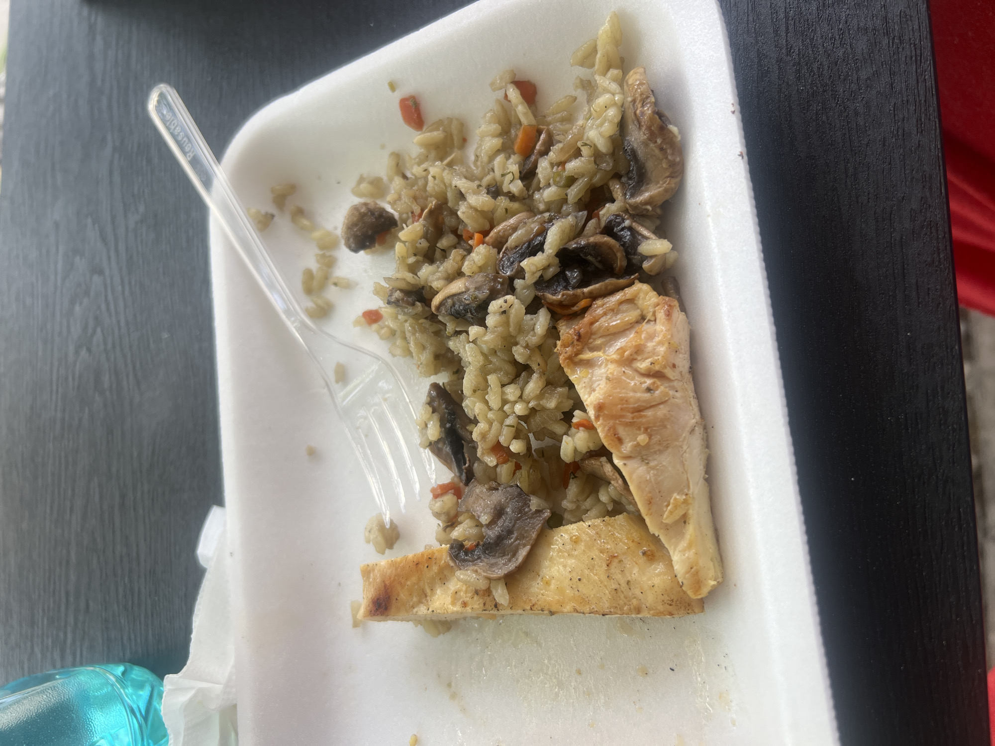 A partially eaten meal sits on a white, disposable styrofoam plate. The meal consists of rice mixed with chopped vegetables and sliced mushrooms, accompanied by pieces of cooked chicken. A clear plastic fork rests on the rice. The plate is placed on a dark, possibly wooden, surface, potentially a table. In the lower-left corner, part of a light blue object is visible. Red is vaguely seen in the background. The picture was most likely taken in Sofia, Bulgaria.