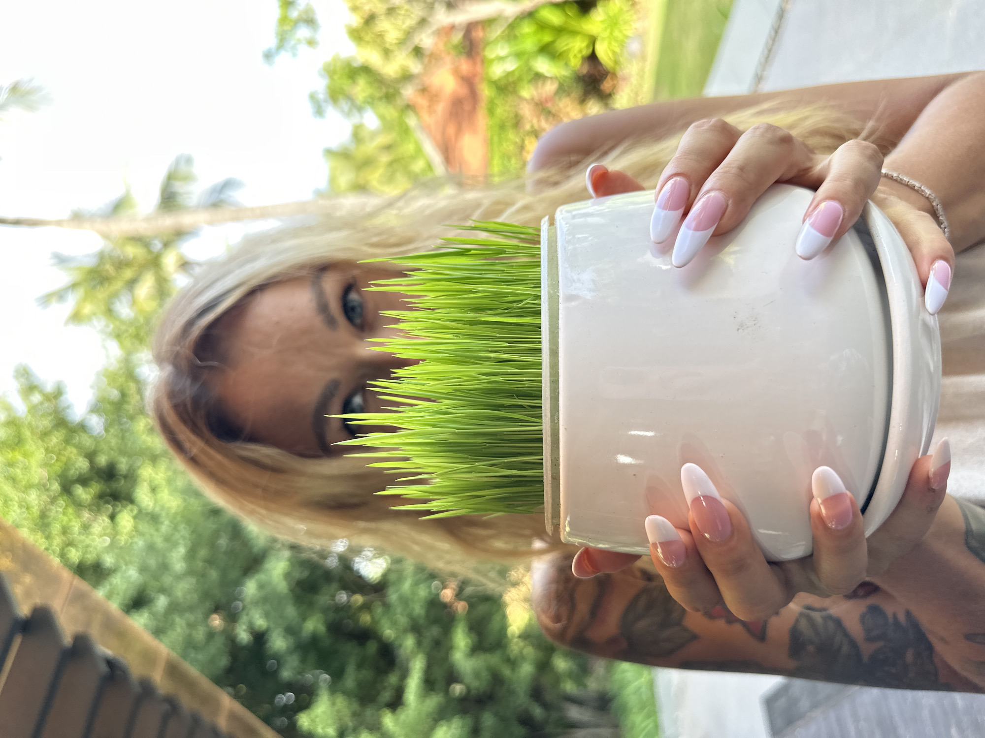 A woman with blonde hair and blue eyes is holding a white potted plant filled with bright green grass in front of her face. She is wearing a light-colored top and has long, almond-shaped nails with a French manicure. Her left arm and shoulder are visible, showing a tattoo. The background is blurred, suggesting an outdoor setting with lush greenery and palm trees, indicative of a tropical or sub-tropical environment. The lighting suggests it is daytime. The location is specified as Phu Thuy Ward, Vietnam.