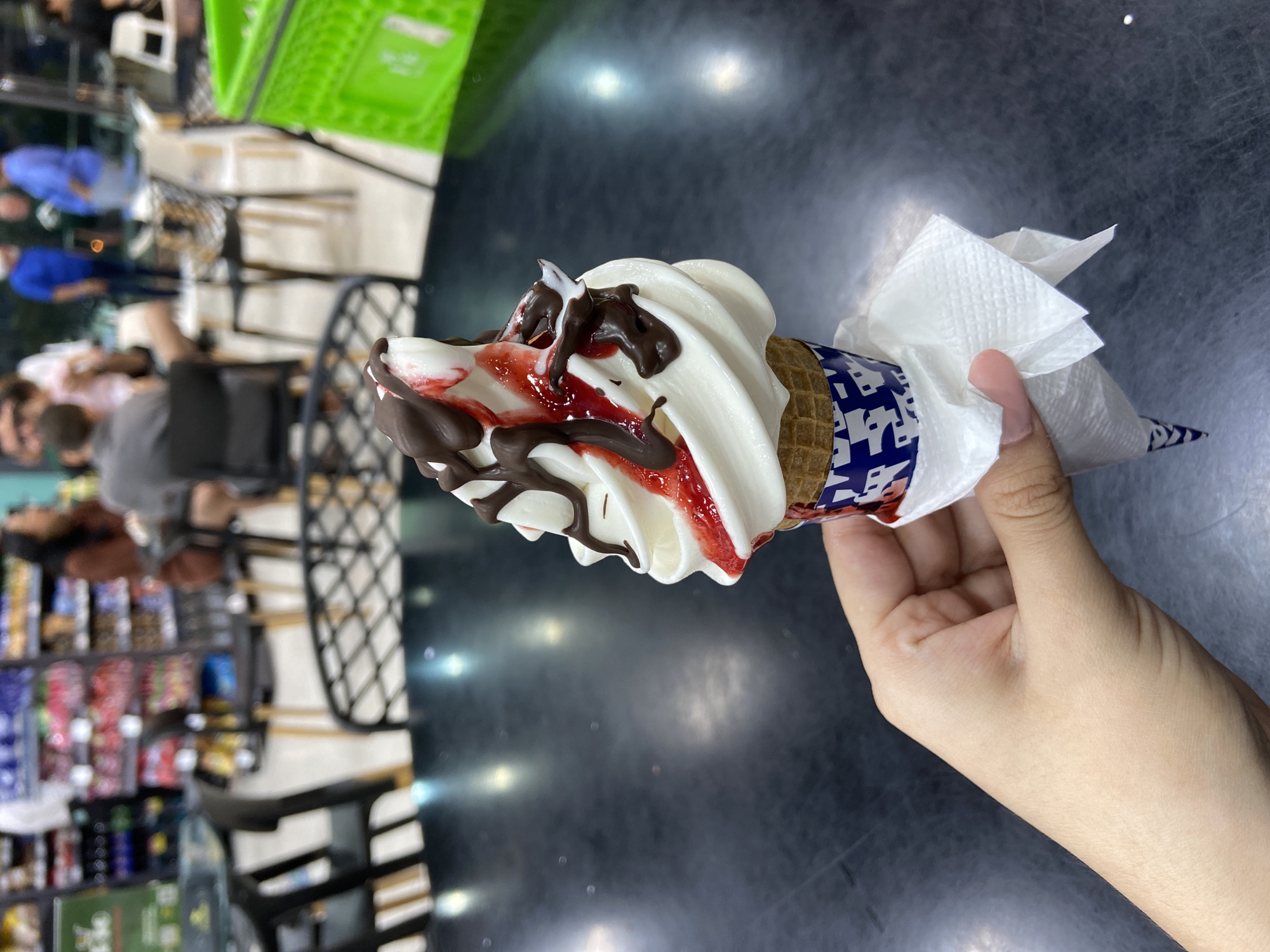 A person holds an ice cream cone with vanilla soft serve, strawberry, and chocolate sauce in their right hand. The cone is wrapped with a white paper napkin. The background shows a blurred indoor scene with supermarket shelves and people. The reflection on the table shows the supermarket details. This photograph was taken in Juan Griego, Venezuela.