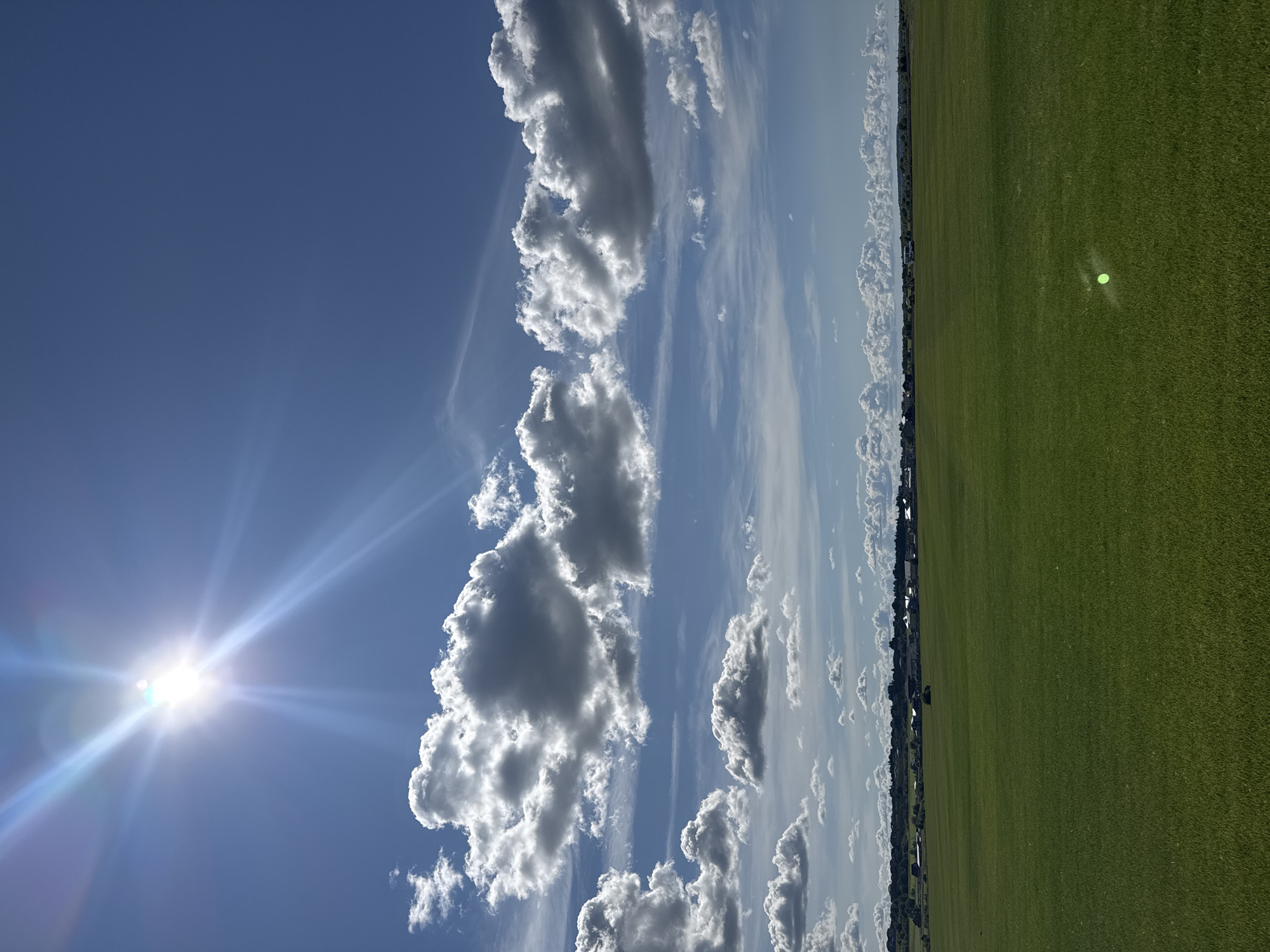 The image captures a wide, sun-drenched landscape in Kempelevo, Russia, characterized by an expansive green field under a vibrant blue sky dotted with fluffy white clouds. The sun is prominently positioned in the upper left quadrant, emitting bright rays and lens flare. A low-lying line of buildings and trees stretches across the horizon in the mid-ground. In the foreground, a bright green circular reflection is visible on the grassy field. The scene is devoid of any people or immediate action, presenting a serene, rural environment.